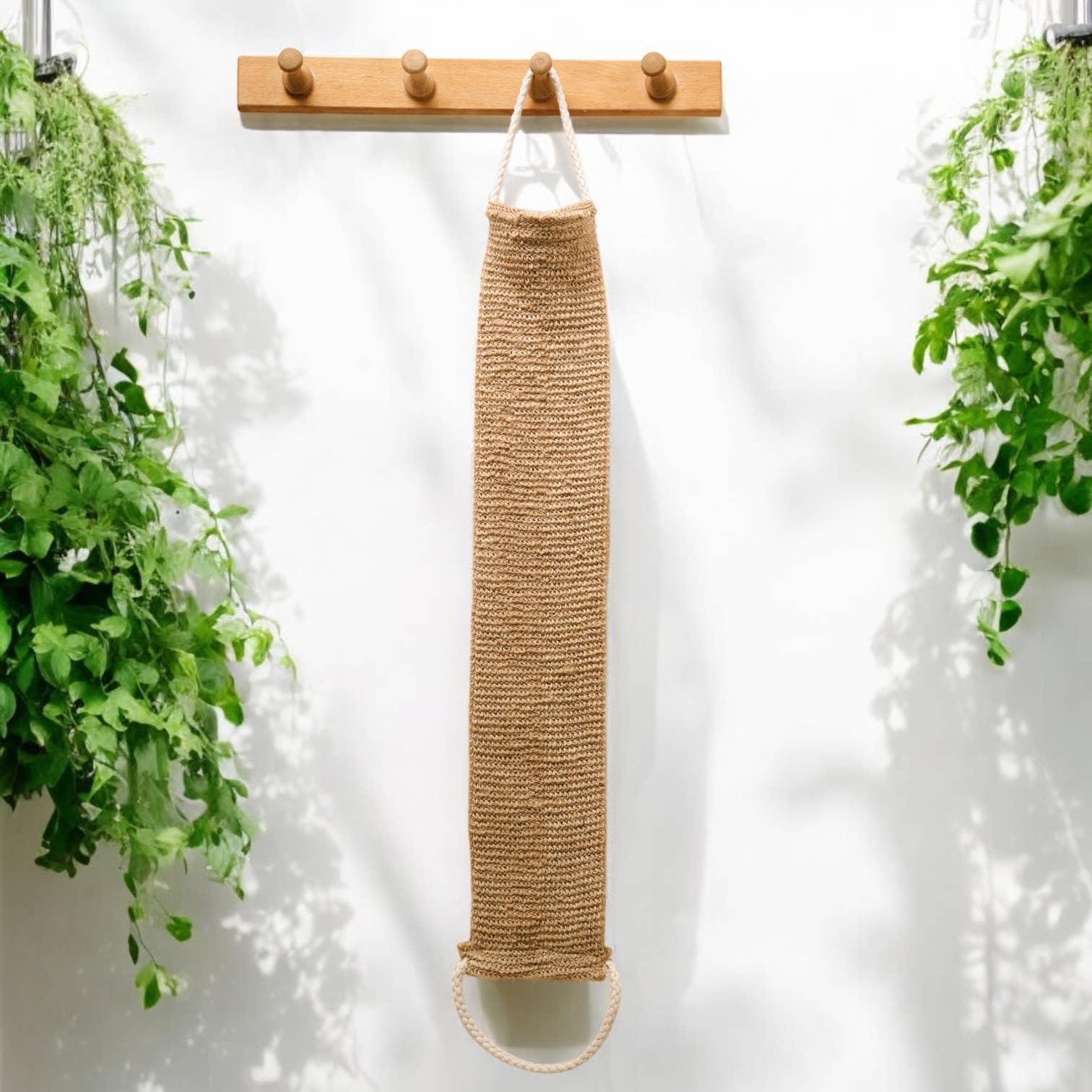 A long jute bag hangs from a wooden rack with five hooks, next to the Scrub: Sisal Linen Back Exfoliating Strap, surrounded by green leafy plants.