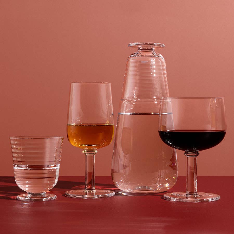 A KostaBoda Viva All Purpose Glass Medium (from the 2-Pack) filled with a golden liquid is arranged alongside a glass carafe and two water glasses on a red surface with a matching background.