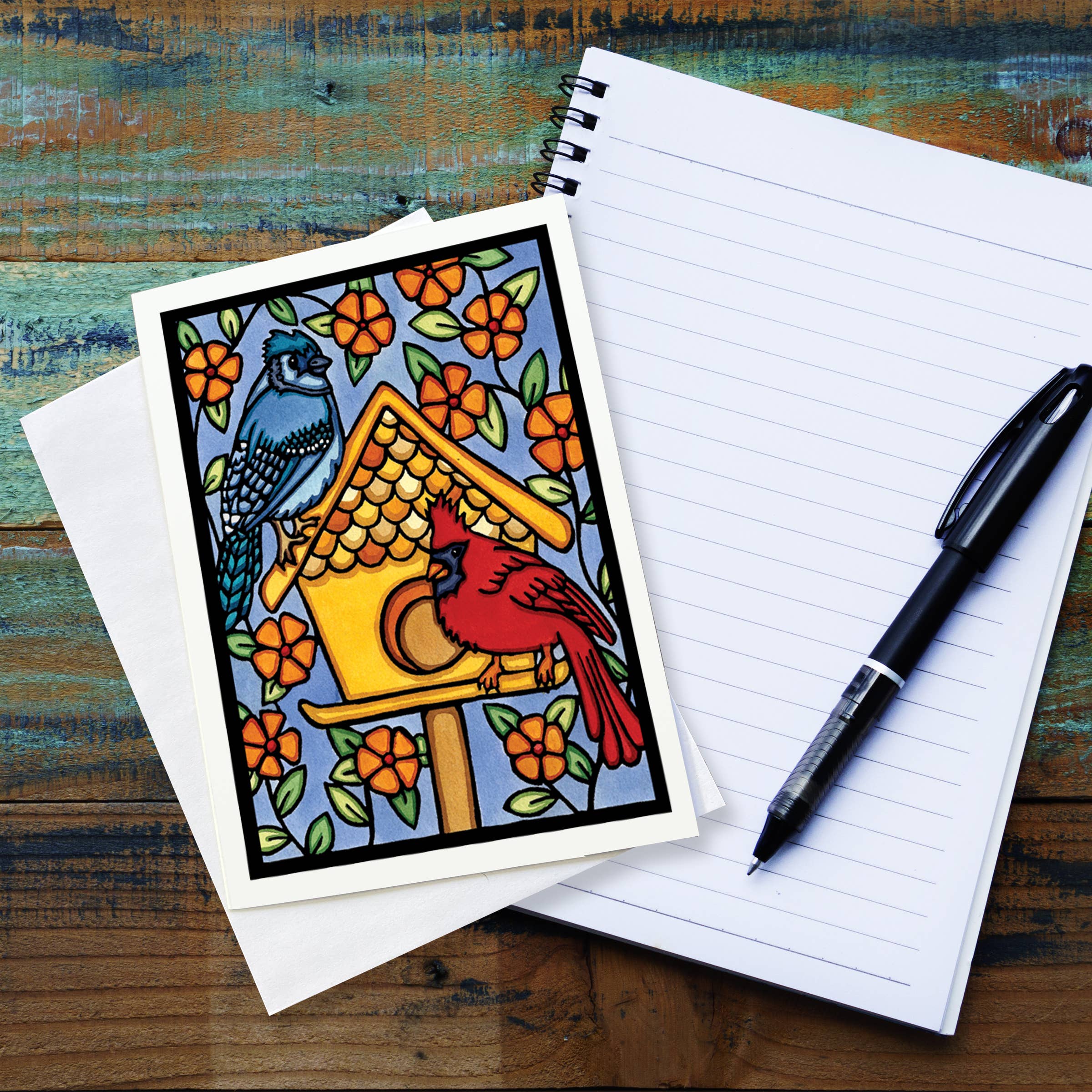 The Card: Birdhouse 5x7 Greeting Card by Sarah Angst features two birds and a birdhouse, comes with an envelope, is blank inside, and is made in America. Shown beside a spiral notebook and pen on a wooden table.
