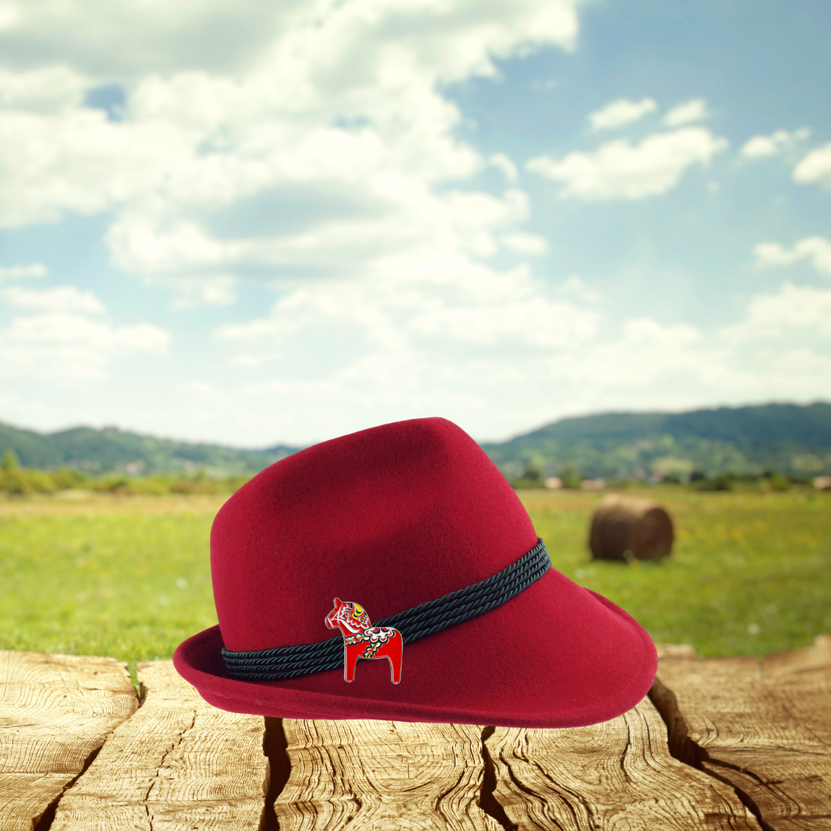 A red hat featuring a Swedish Dala Horse metal pin rests on wooden planks in a field, with hills and cloudy skies behind it.