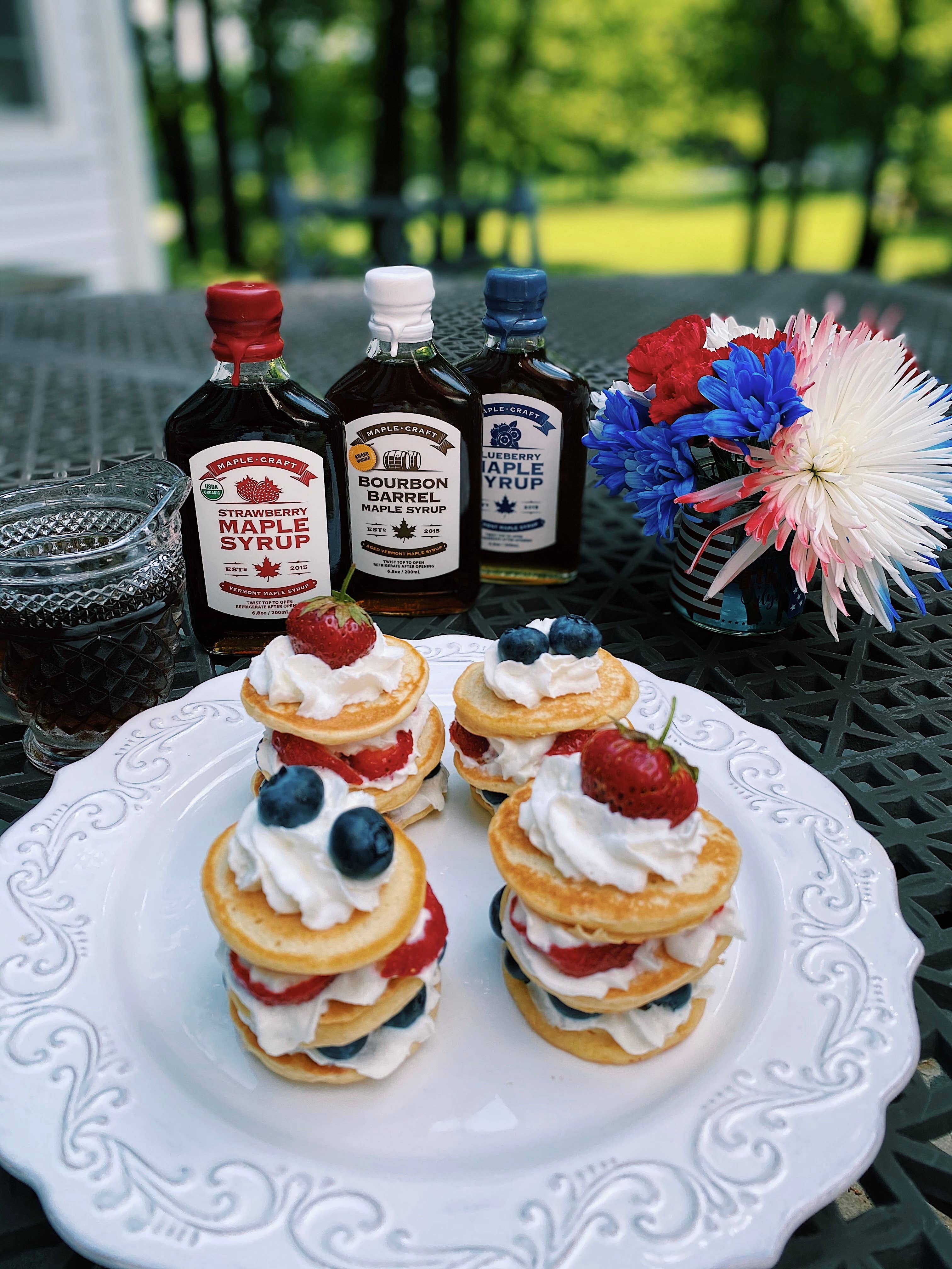 Three small pancake stacks with fruit and cream sit on a white plate. In the background, Summer Favorites Strawberry Maple Syrup bottles and a floral centerpiece decorate a metal table.