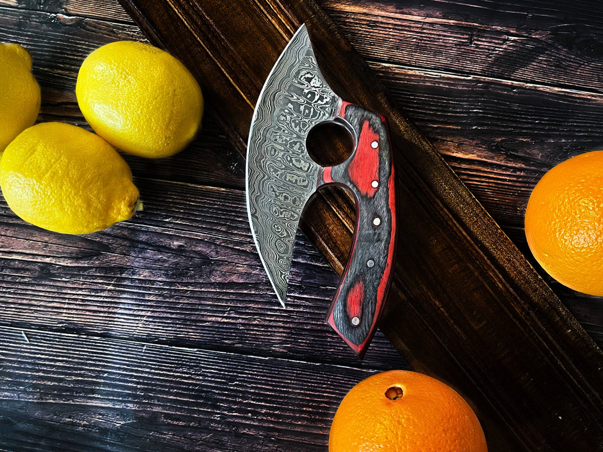 An Alaskan Ulu Style Blade kitchen knife with a red and black diamond wood handle rests on a wooden surface beside three lemons and two oranges. The handmade blade is crafted from Damascus steel.