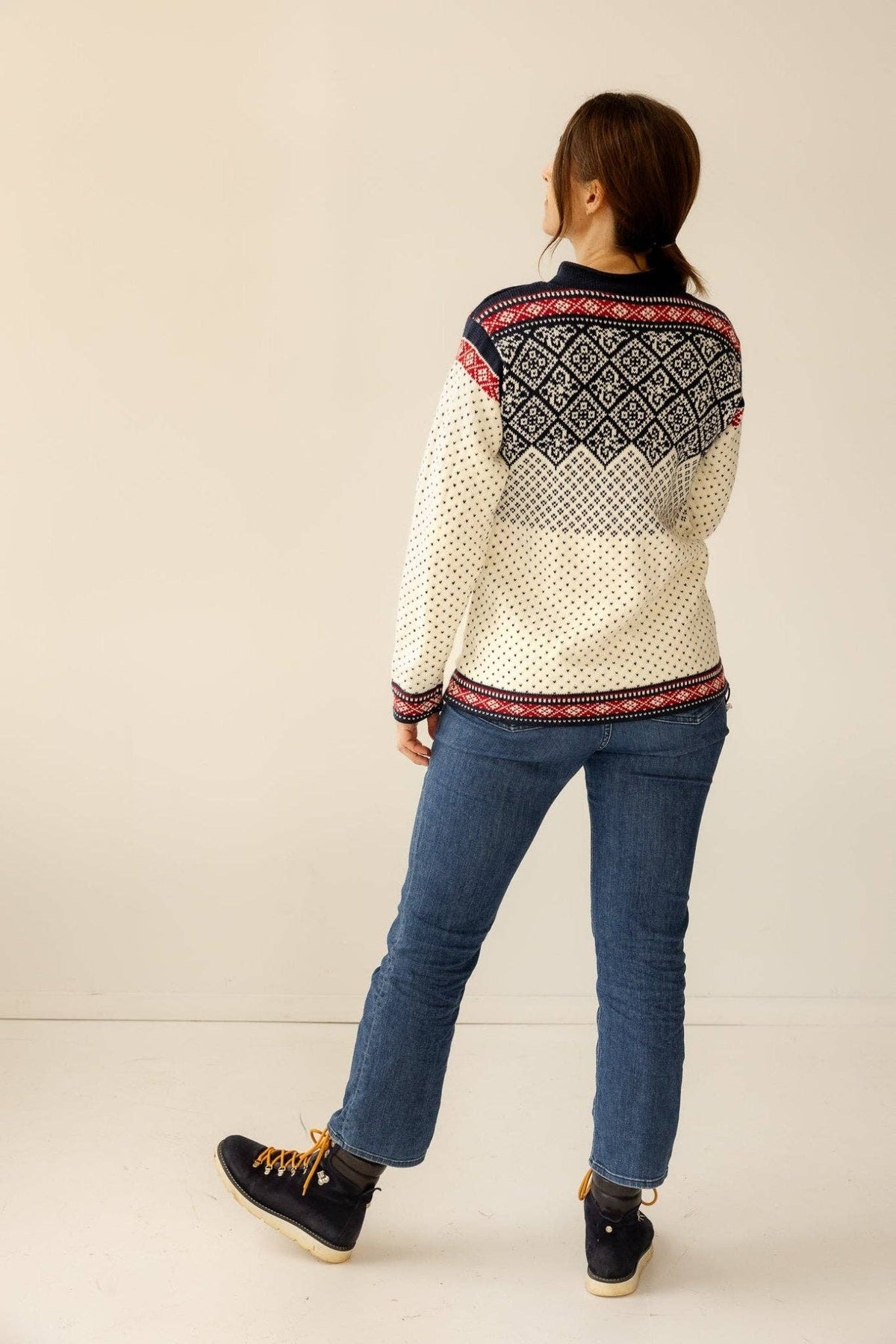 Off-white Norwegian wool cardigan featuring dark blue geometric patterns and red accents, worn with blue jeans and dark boots.
