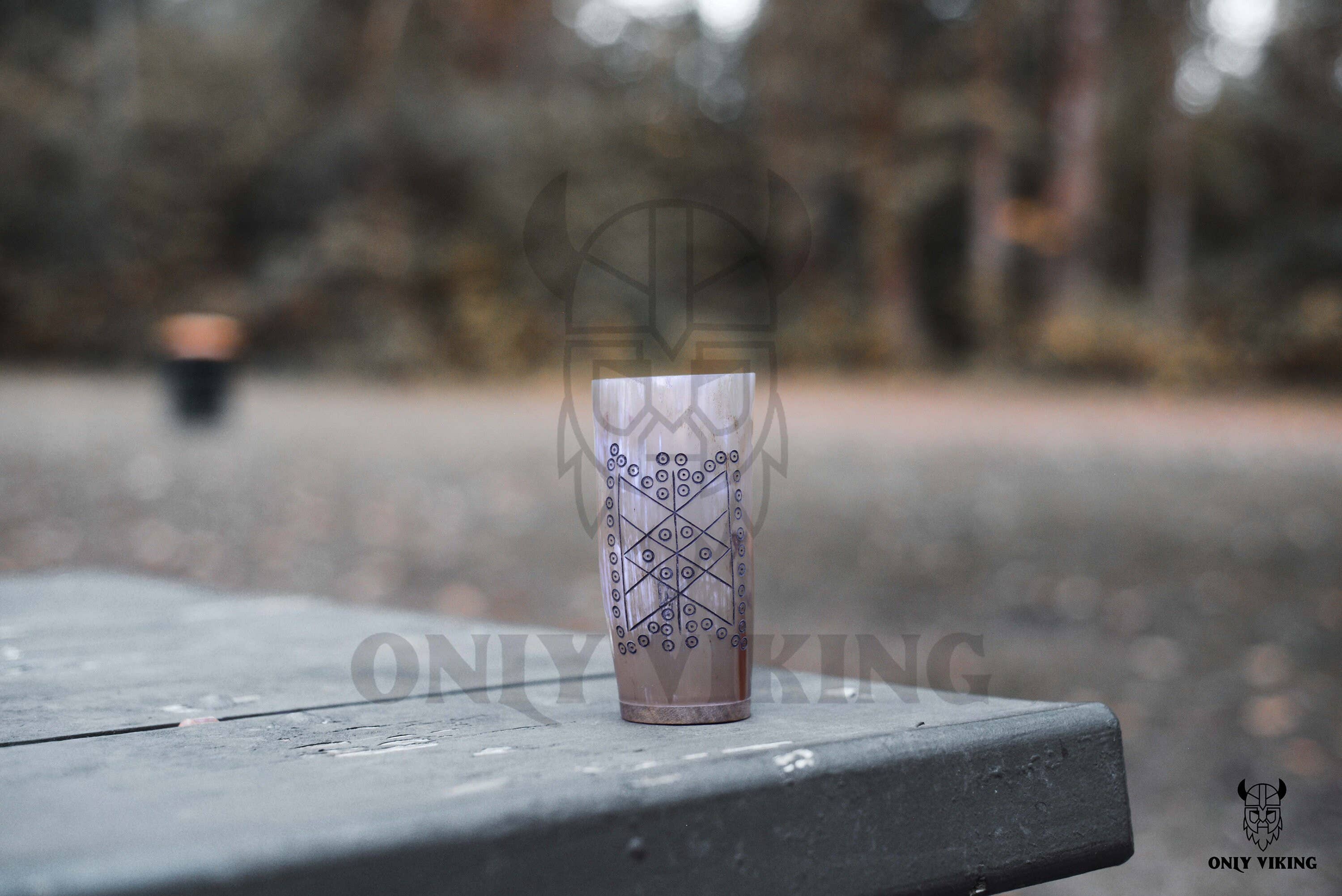 A Horn Mug: Beer Stein Drinking Horn Mug Tree of Life (6-9 oz) with geometric patterns rests on a wooden outdoor table, with blurred trees and a trash can in the background.