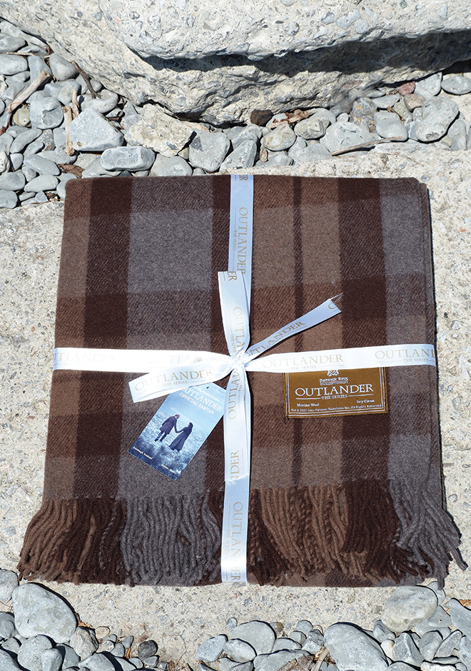 A folded brown Blanket: Outlander Merino Wool Blanket with fringed edges rests on rocks, tied with a white ribbon and an Outlander-themed tag.