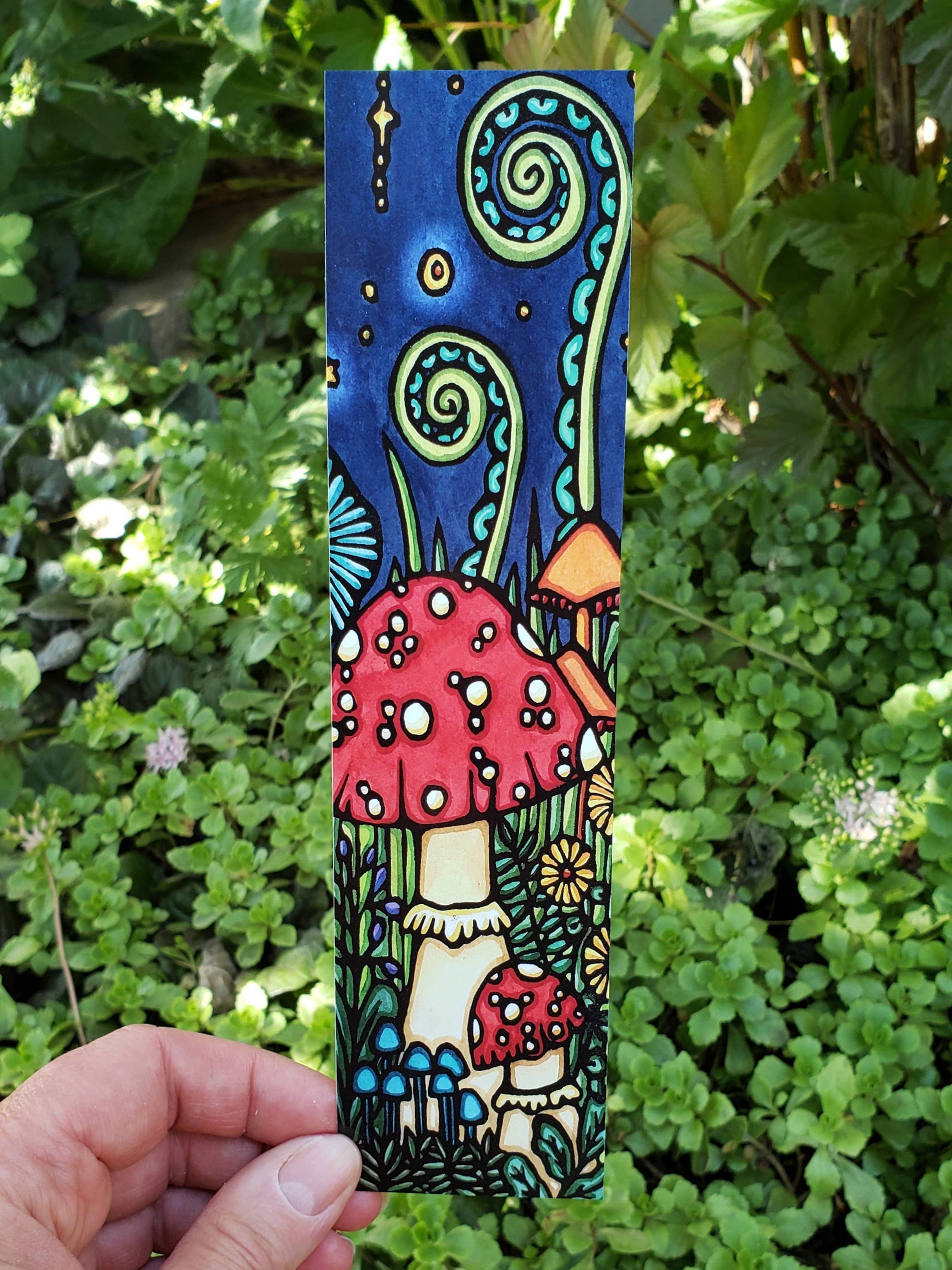 A vibrant bookmark titled "Mushroom" by Sarah Angst, featuring hand-drawn red mushrooms, stars, and swirling patterns printed on heavy cardstock with a matte gloss finish, displayed against a backdrop of green foliage.