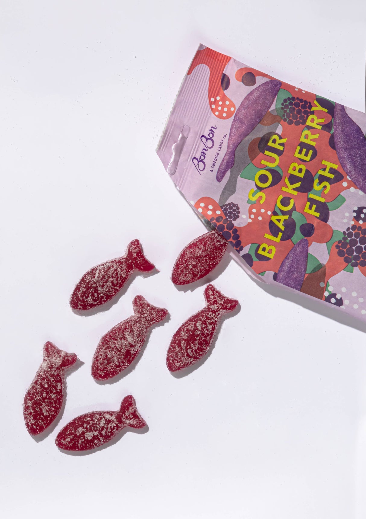 An opened package of Candy: Bon Bon - Sour Blackberry Fish (150g) 5.2oz displays six red, sugar-coated vegan fish-shaped gummies scattered outside—a fun twist on classic Swedish candy.