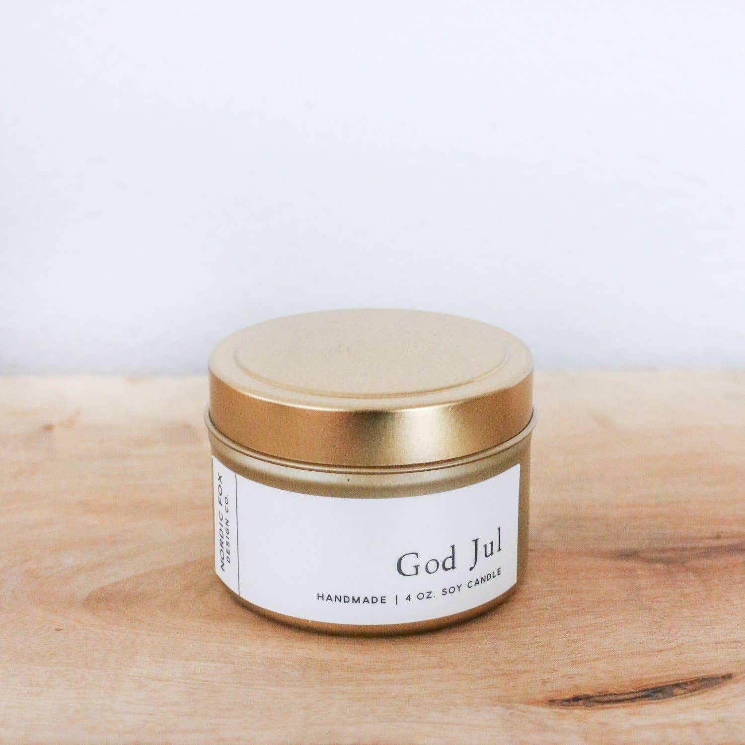 A 4 oz. God Jul Handmade Soy Candle in a small gold tin with a white label rests on a light wooden surface, filling the air with a festive holiday scent.