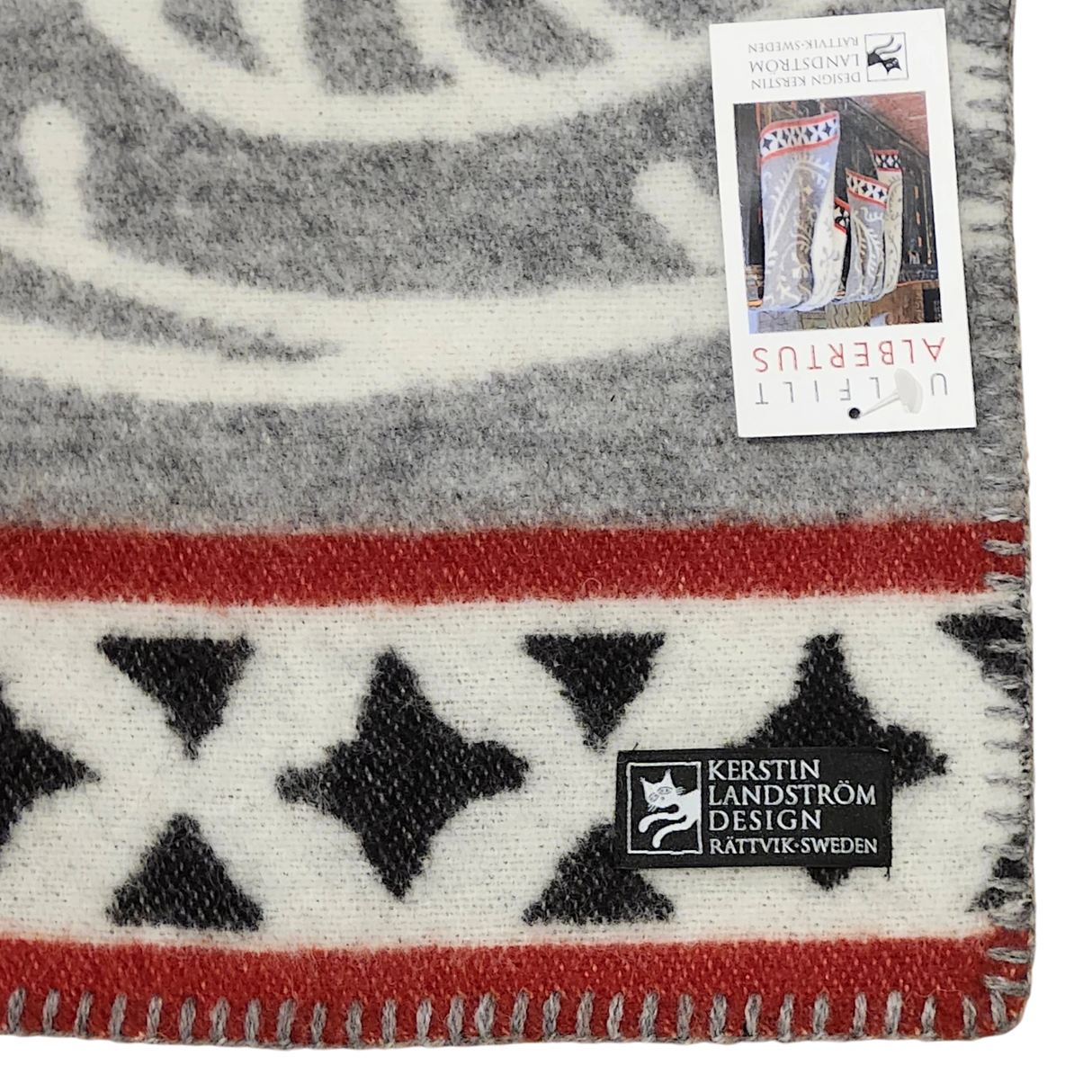 Close-up of a Swedish wool blanket featuring black stars, red and gray stripes, a stitched edge, and two labels, one marked Kerstin Landström Design.