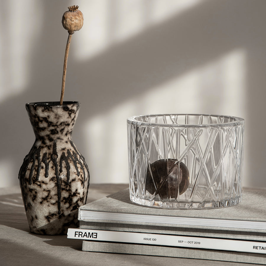 An Orrefors City Bowl, designed by Martti Rytkönen, cradles a stone beside a textured vase with a dried flower, all elegantly situated on stacked magazines under soft lighting for a tranquil scene.