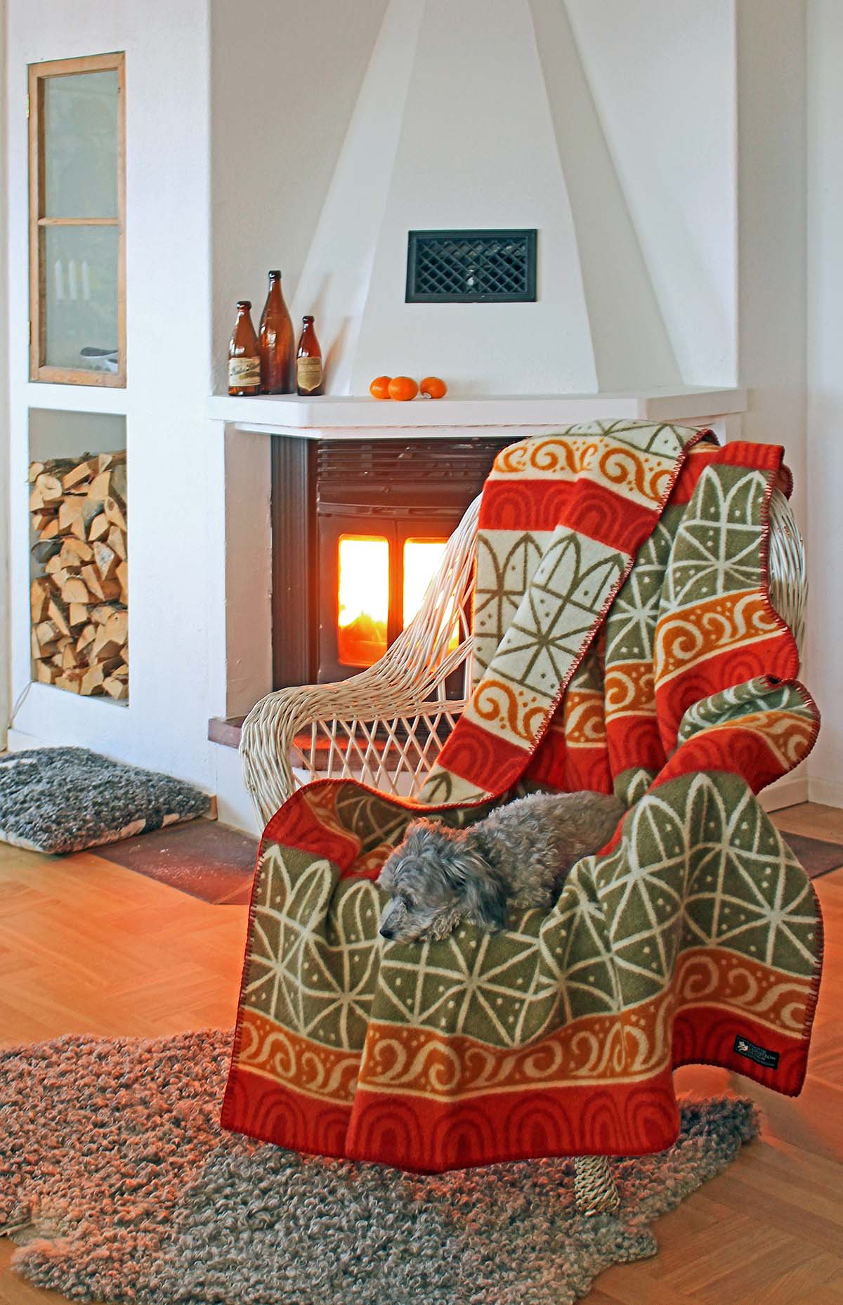 A cozy room features a wicker chair with the Uppsala - 100% Norwegian Lambs Wool Blanket by Kerstin Landstrom draped over it, where a small dog rests. The warm glow from the fireplace, paired with neatly stacked firewood and bottles, enhances the inviting ambiance.