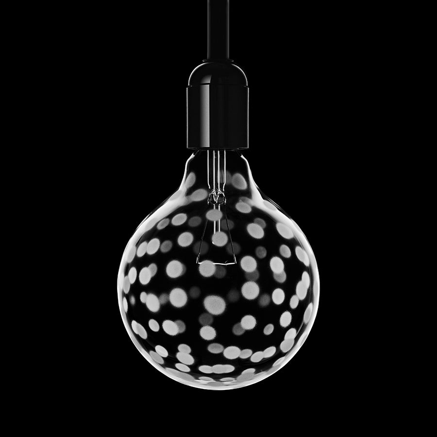 The Orrefors: Light Shadow Confetti 25W Light Bulb features sand-blasted dot motifs, suspended against a black background for a minimalist design.