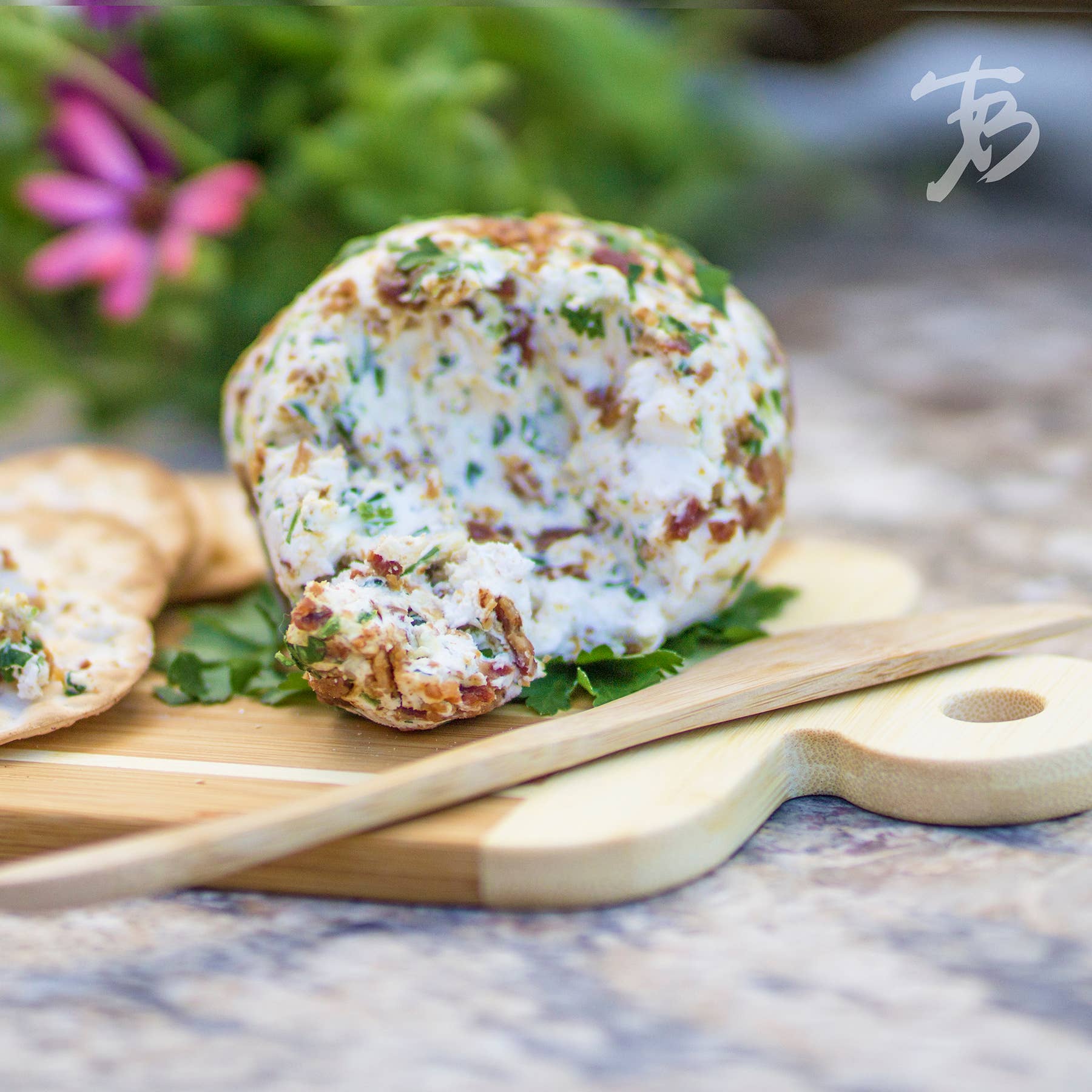 A striped paddle serving and cutting board, accompanied by a spreader knife, displays an herb and spice cheese ball next to crackers. Flowers are blurred in the background.