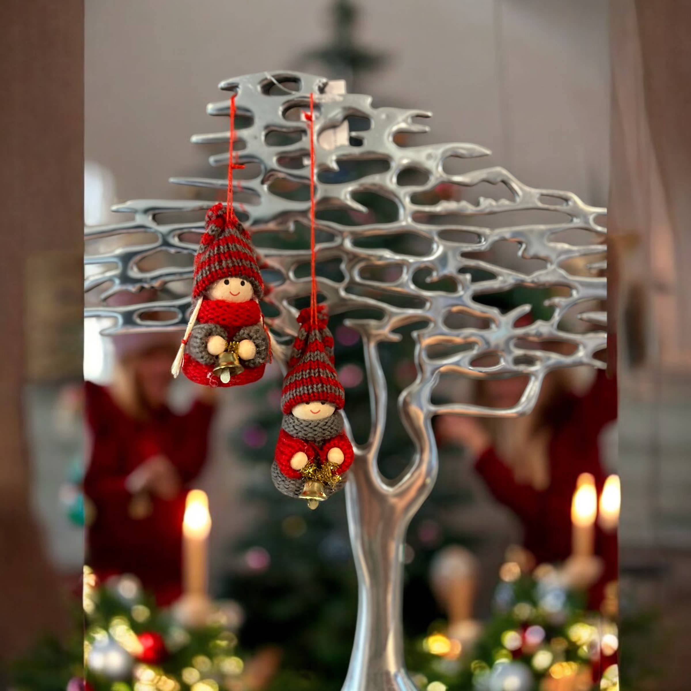 Two Tomte Boy or Girl with Bells Christmas Tree Ornaments hang on a silver metal tree sculpture, surrounded by candles and greenery, with a blurred Christmas tree in the background—ideal for Scandinavian holiday décor.