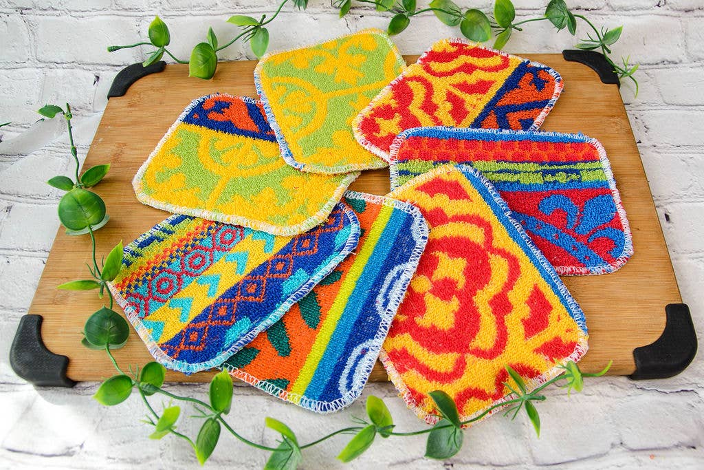 Several colorful, patterned Scrub: Kitchen Scrubber Scrub-Wow coasters are arranged on a wooden board with green vines, set against a white brick background. Each is made from multi-purpose, Teflon-safe, non-toxic scrubby material.