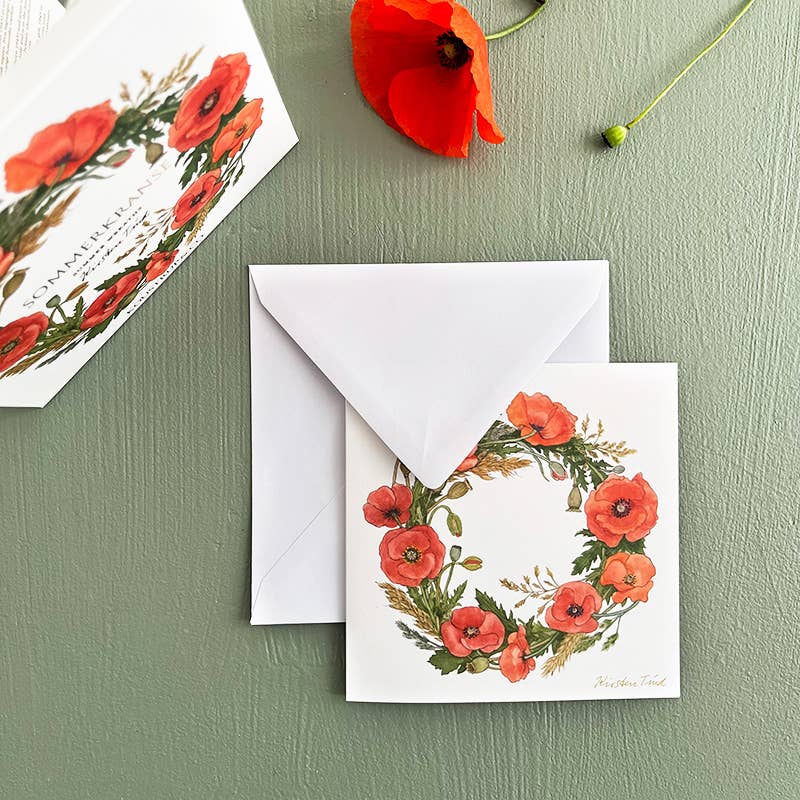 A charming square card folder from Koustrup & Cos Card Pack: Summer Wreaths, featuring 8 unique designs of poppy flower wreaths, adorns an envelope. Nearby, a vibrant red poppy on green captures the essence of its elegant Danish design.