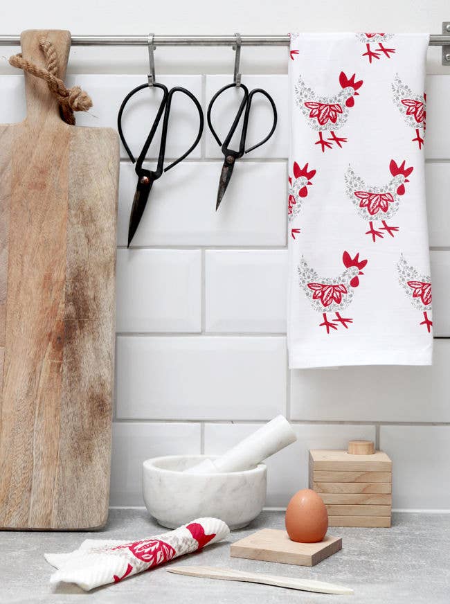 In a cozy kitchen scene, two pairs of scissors rest by a cutting board. The charm is elevated by the Hen Tea Towel - Grey & Red, featuring Scandinavian-designed cotton with red chicken patterns. Nearby are a mortar and pestle, stacked wooden blocks, a brown egg, and a vibrant cloth.