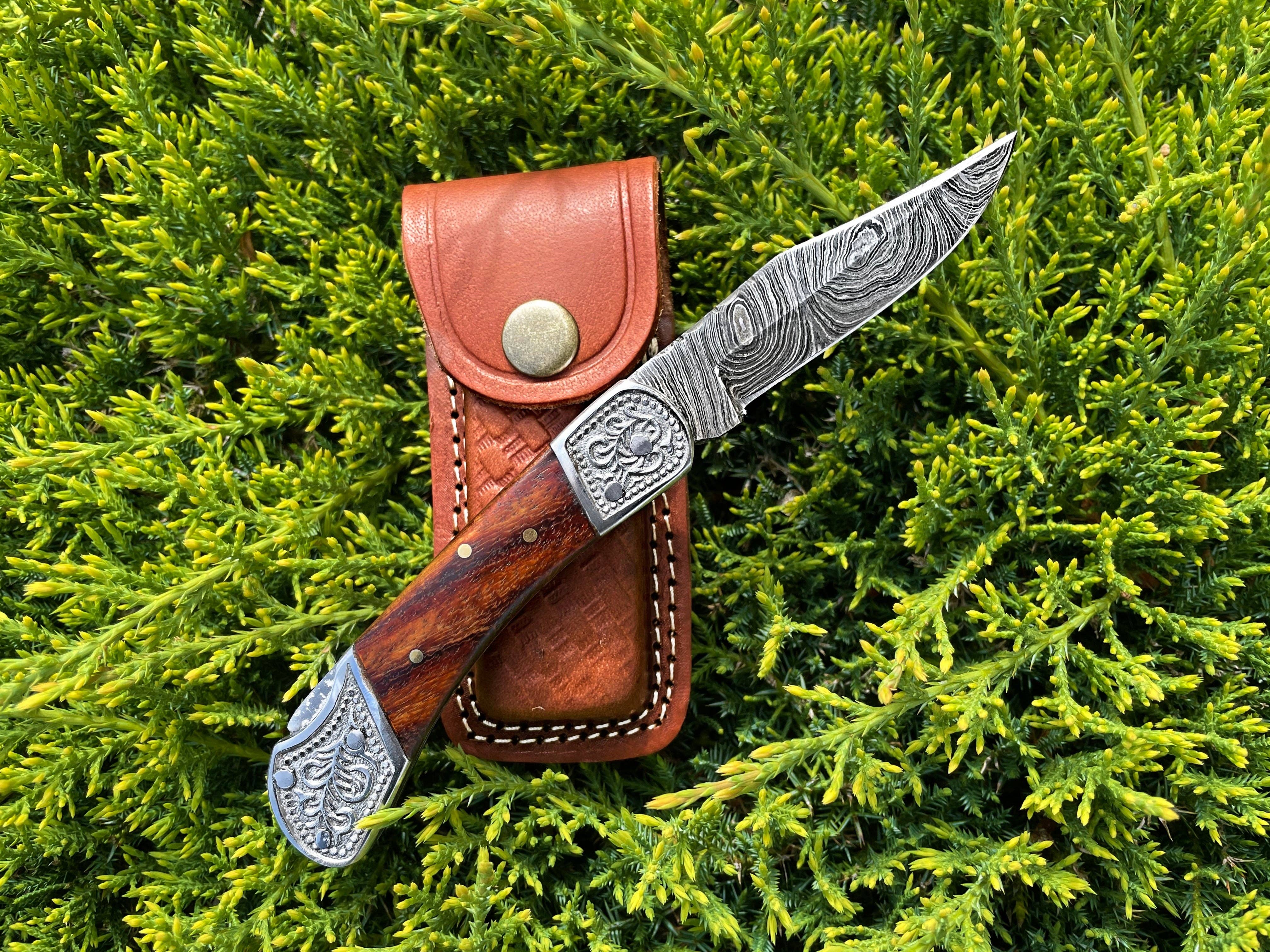 The Pocket Knife Walnut Wood 6.5'' Damascus Steel, featuring engraved metal, rests atop a brown leather pouch on green foliage.