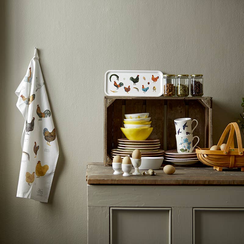 A kitchen counter features stacked dishes, yellow bowls, eggs, and a wooden basket. A wall-mounted rack holds a chicken-patterned towel. A wooden crate showcases the Colorful Hens on Birch Veneer serving tray and containers, adding charm to the scene.