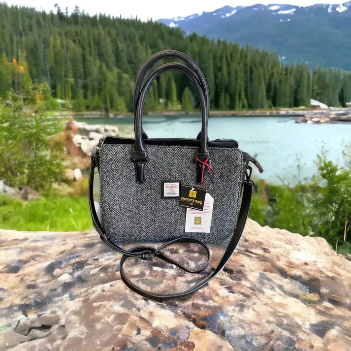 The Bag: Harris Tweed Double Handle Tote - Grey rests on a rock, its black handles and tags contrasting with the scenic backdrop of a lake, trees, and mountains.