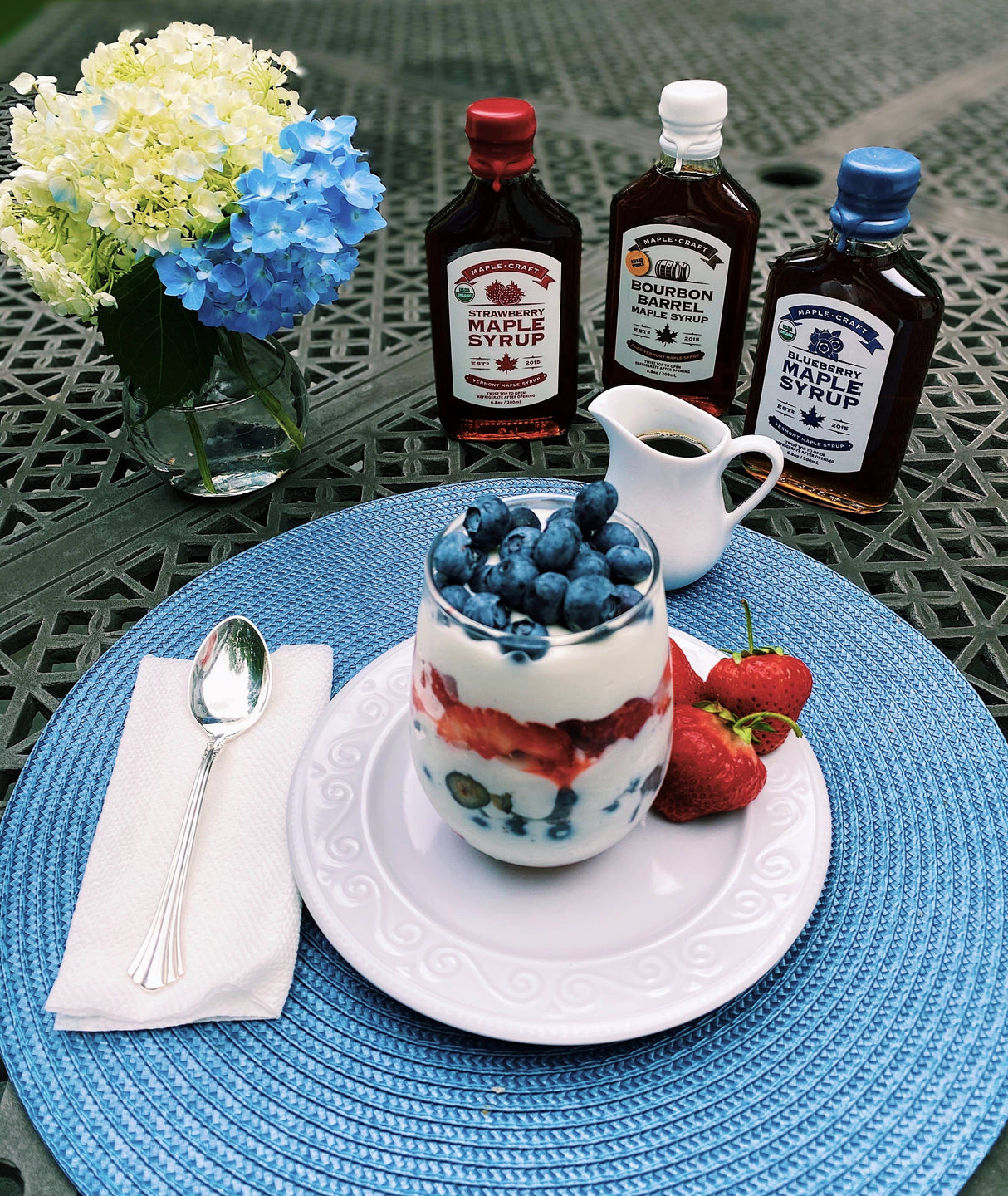 A parfait with blueberries and strawberries on a white plate sits on a blue mat, accompanied by three syrup bottles, including Maple Syrup: Strawberry - Summer Favorites Maple Syrup (6.8 oz). A small pitcher, spoon, and flower arrangement complete the refined sugar-free setting.