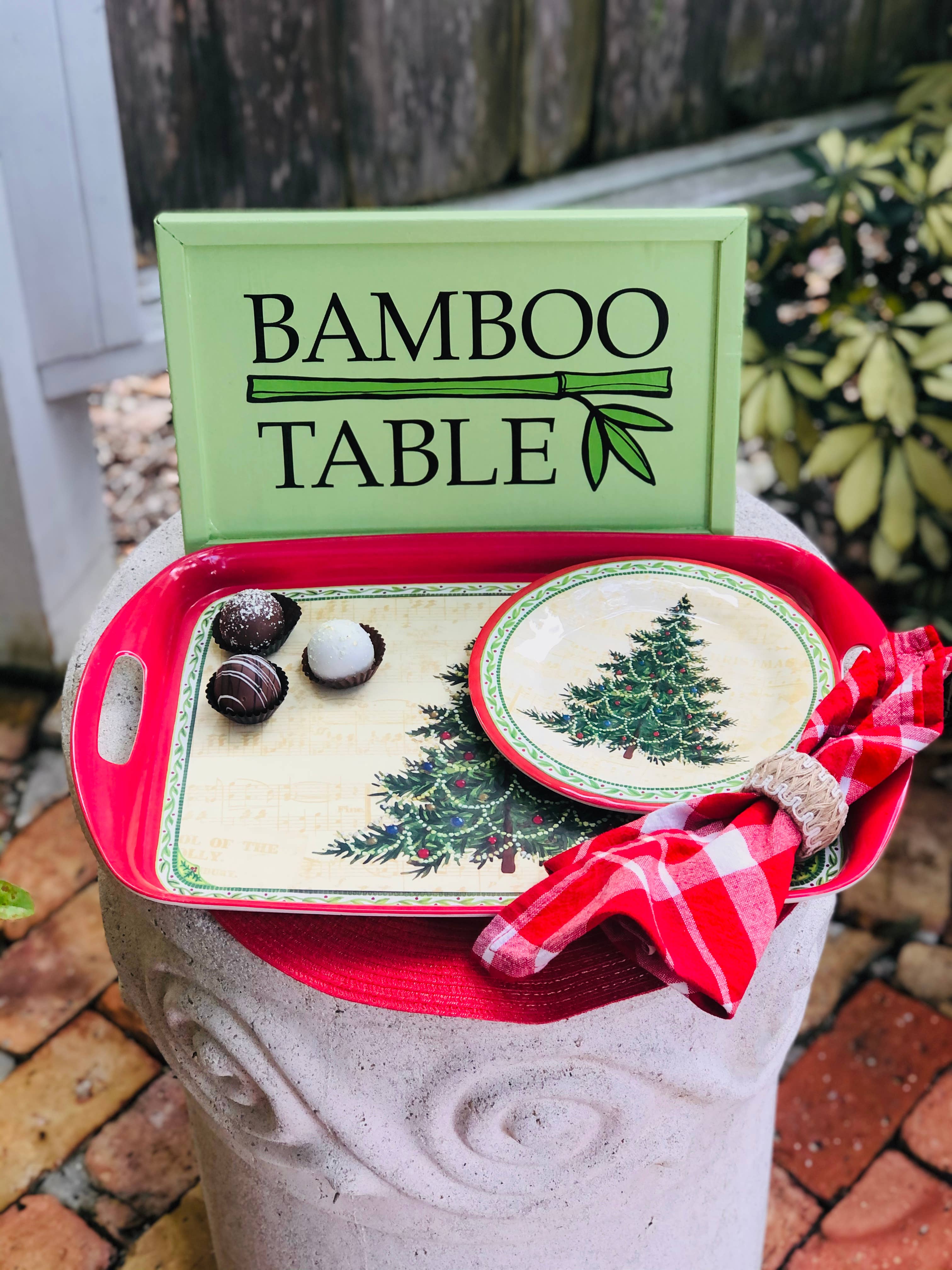 The Merry Christmas Tree Bamboo Serving Tray sits with Christmas plates, a red checkered napkin, and chocolates in front of a green BAMBOO TABLE sign—an ideal Christmas hostess gift.
