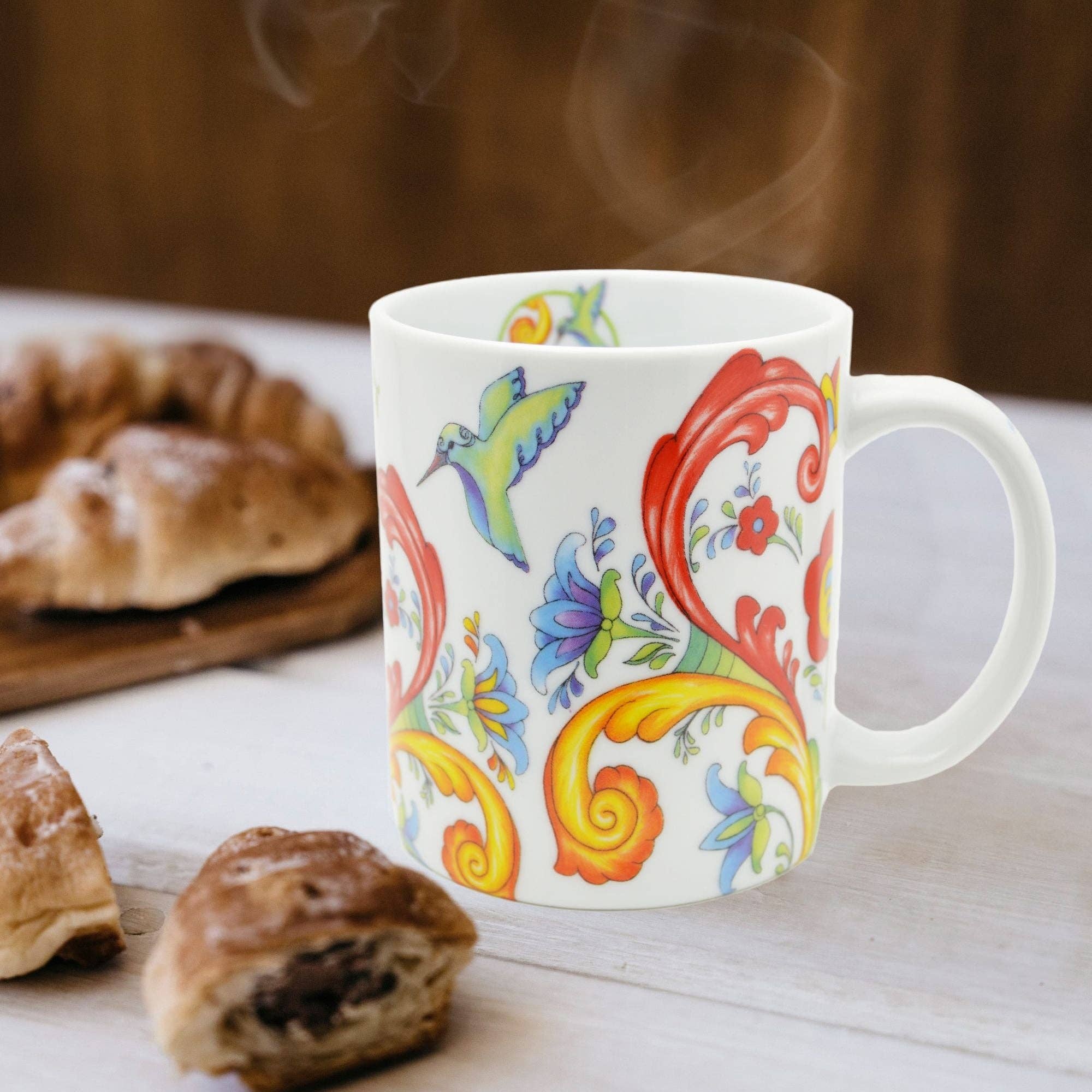 A white 12 oz Mug: Colorful Rosemaling Coffee Mug with vibrant floral and hummingbird artwork sits on a table near pastries, steam rising from its rim.
