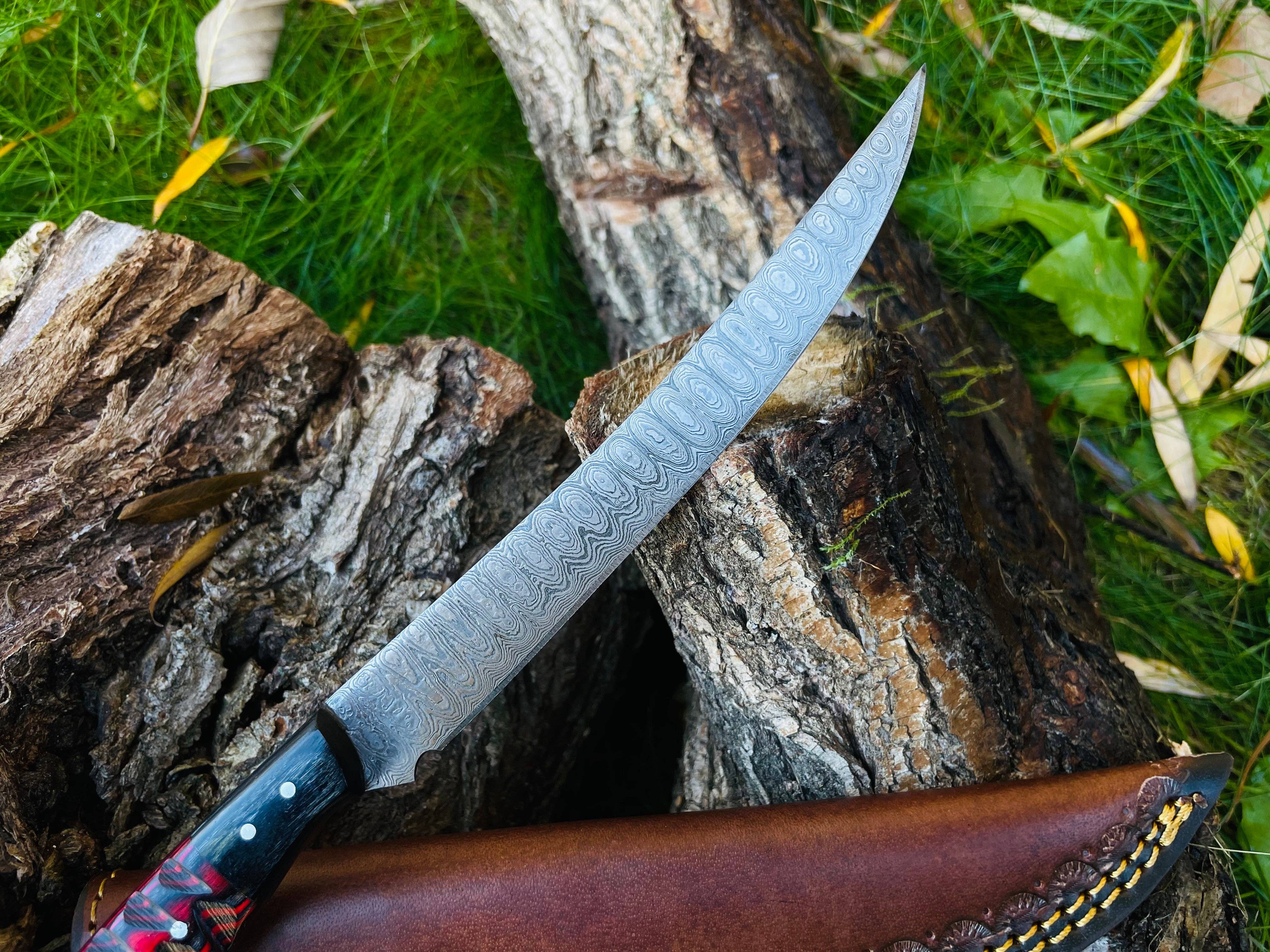The Knife: Damascus Steel Custom Flay Knife with Craved Grips sits on tree logs, leather sheath beside it and green grass behind—an ideal camping knife for outdoor adventures.