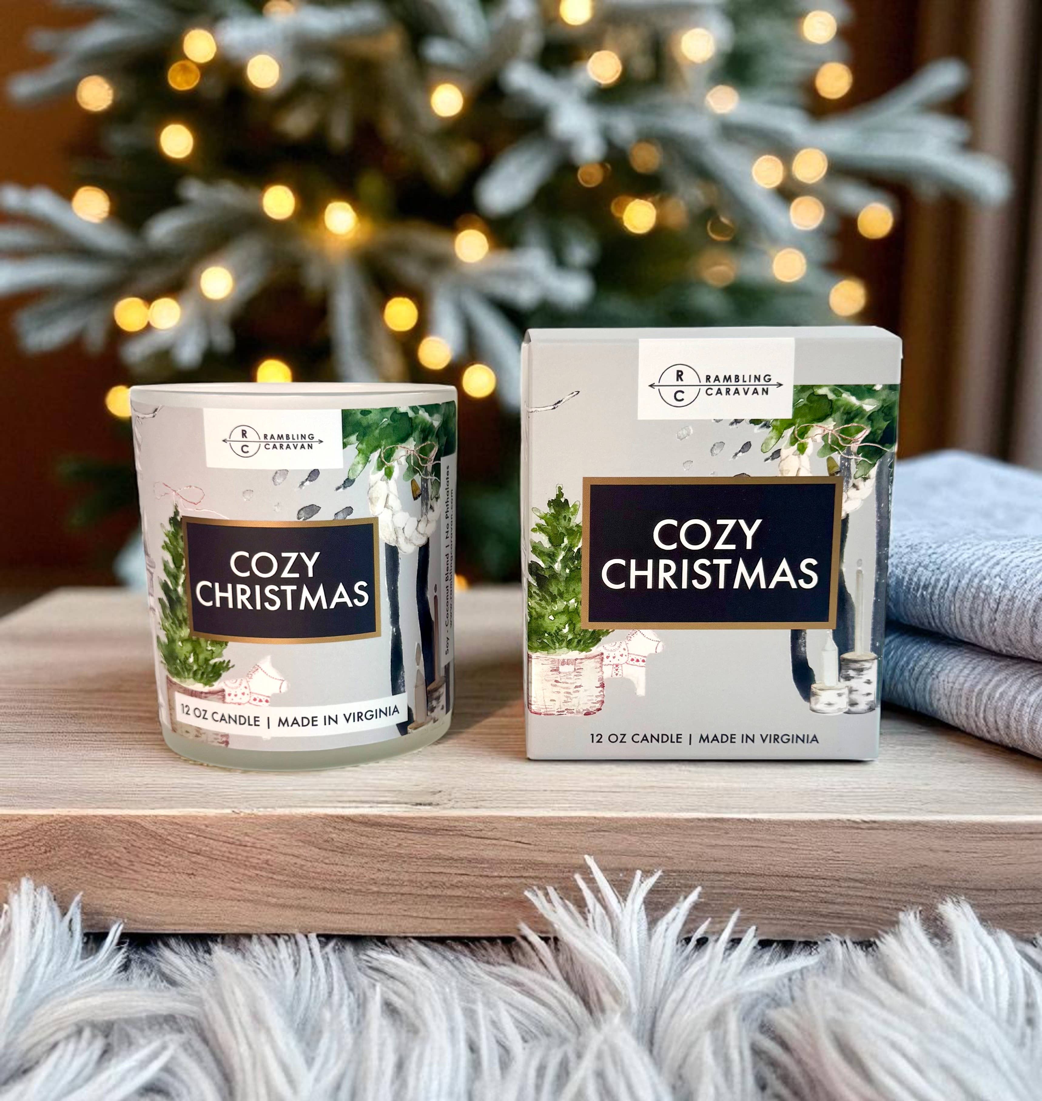 The Cozy Christmas Candle 12 oz and its box sit on a wooden surface with a fuzzy rug, greenery, and a glowing Christmas tree in the background, creating a festive atmosphere with its warm holiday scent.