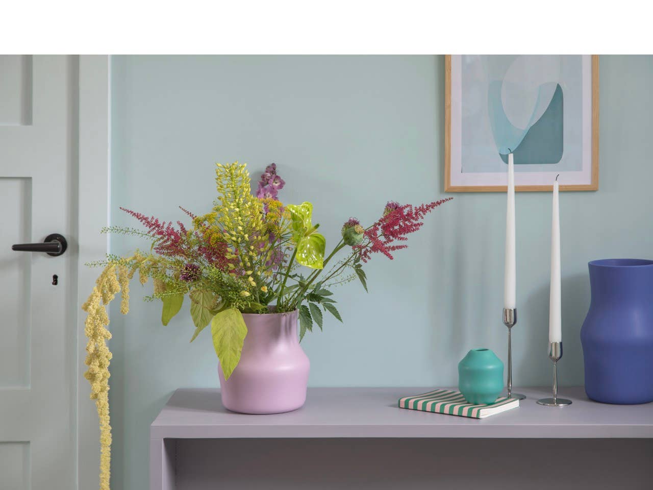 A pink vase with assorted flowers sits on a light gray table next to candles in a Gense Dorotea 2.7 x 7 Polished Steel Candle Holder, a blue Monica Förster vase, a green vase, and a striped notebook, against a pastel-colored wall.