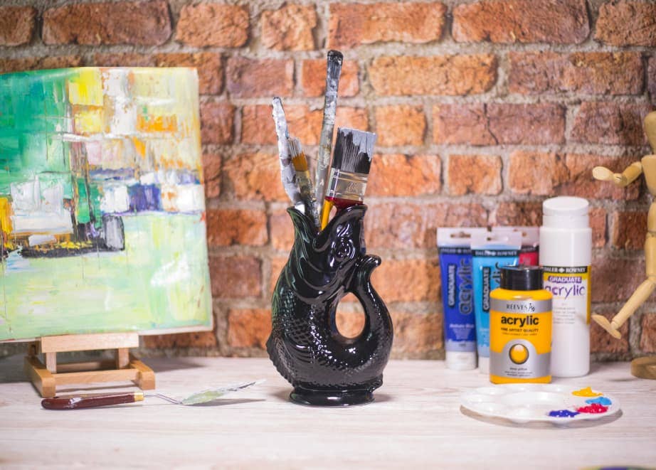 A Gluggle Jug: Black Fish Shaped Pitcher that Gluggles holds paintbrushes on a table with an abstract painting on a small easel, acrylic paints, and an artist’s mannequin, all set against a brick wall.