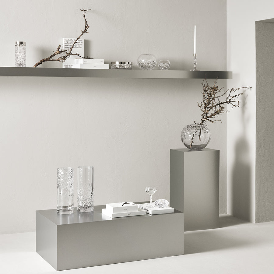 Minimalist interior with gray platforms and shelves displaying clear glassware, books, bare branches in vases, and the Orrefors Carat Candlestick Silver Small Single. Crystal accents enhance the serene elegance against a white wall.
