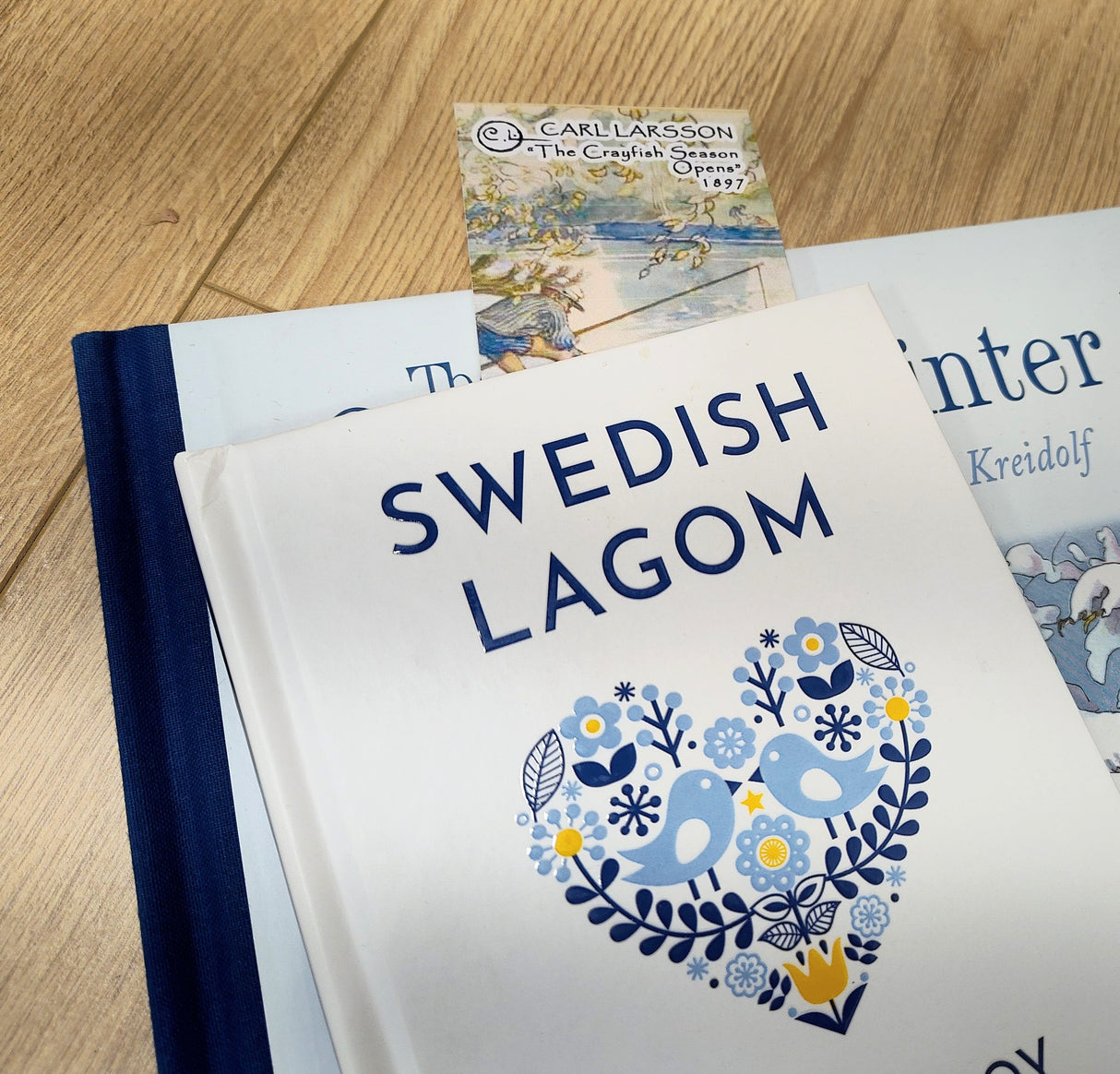 A book titled Swedish Lagom with a blue and yellow heart design rests atop another, with the Bookmark: Carl Larsson “The Crayfish Season Opens” (8.5" × 2.5") partially visible on a wooden surface—ideal for Scandinavian art fans.