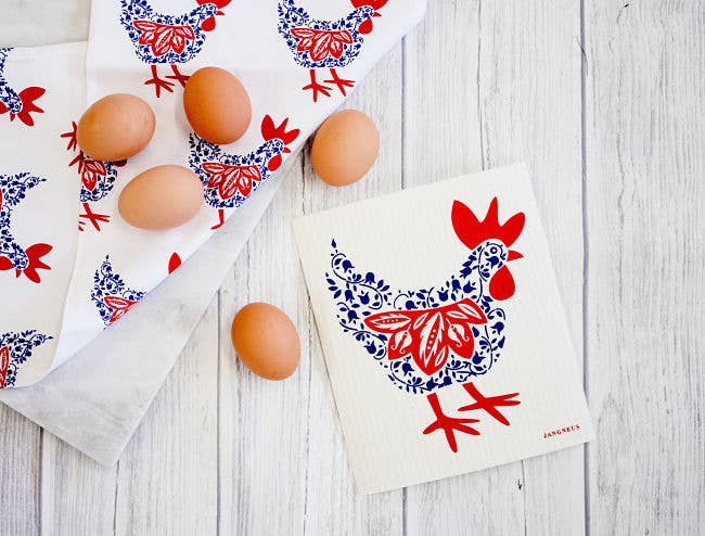 The Swedish Dish Cloth Hen - Blue & Red is artfully displayed on a wooden surface with six brown eggs, accompanied by a matching biodegradable cloth adorned with the same charming chicken motif.