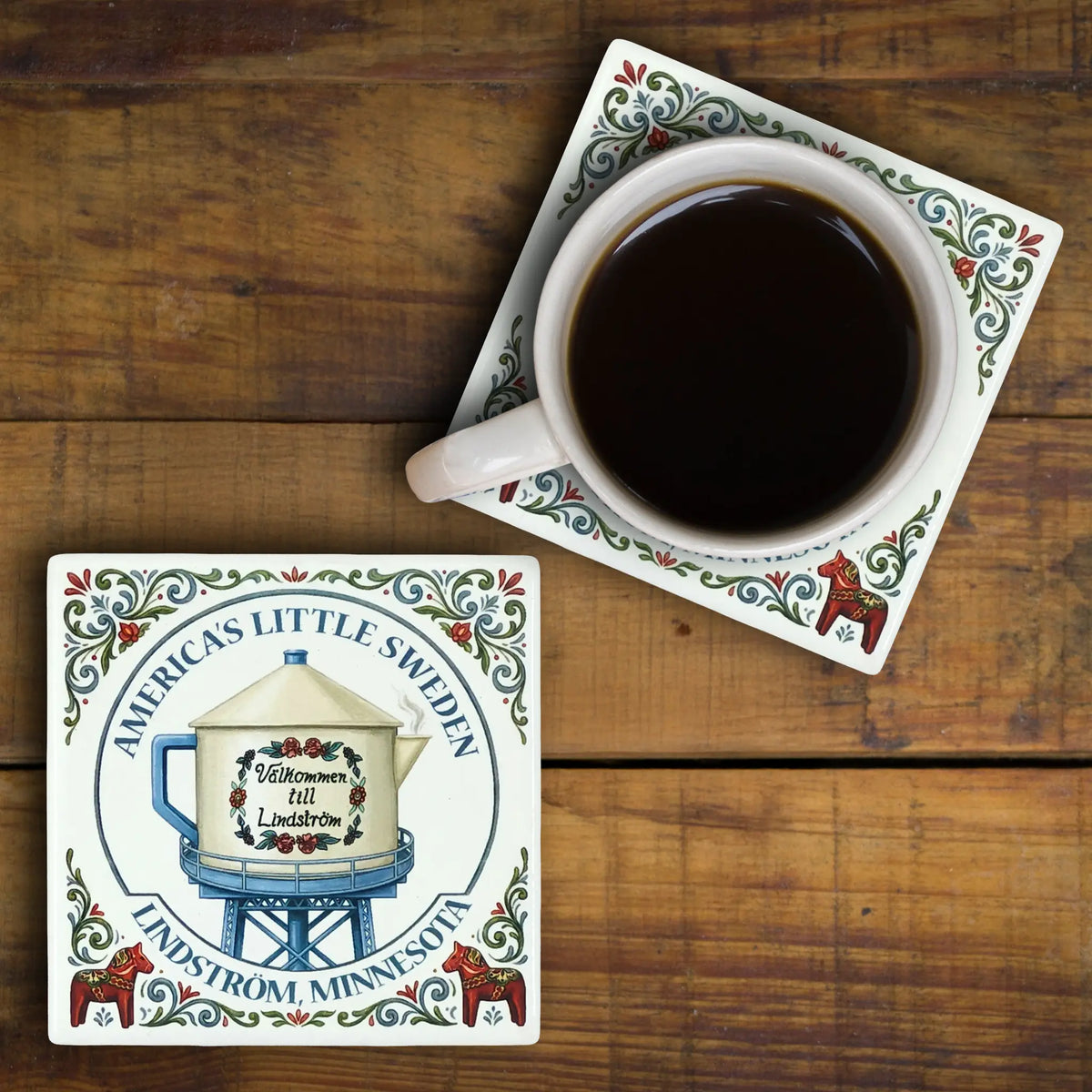 Two ceramic coasters featuring rosemaling and Dala horse designs, one with an illustration of the Lindström water tower and a mug of black coffee.