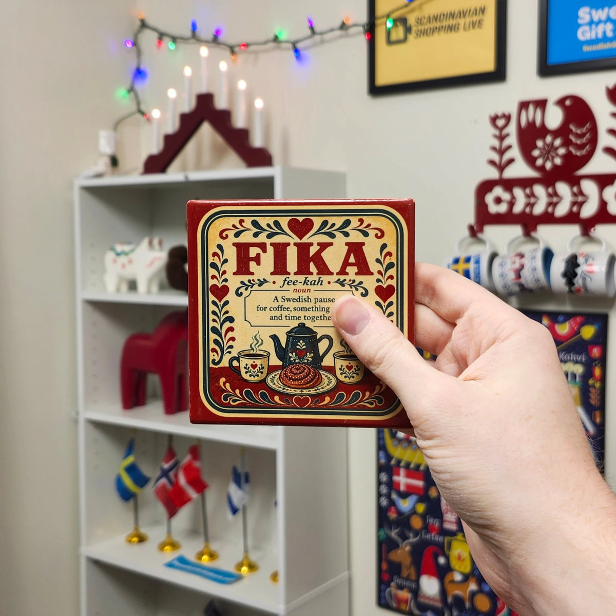 A hand holds a square Swedish Fika coaster featuring the word FIKA, a teapot, cinnamon buns, and floral illustrations.