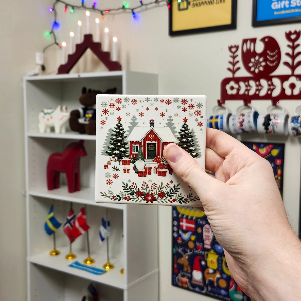 A hand holds a square Swedish folk art coaster featuring a red house, pine trees, and a tomte gnome surrounded by wrapped gifts.