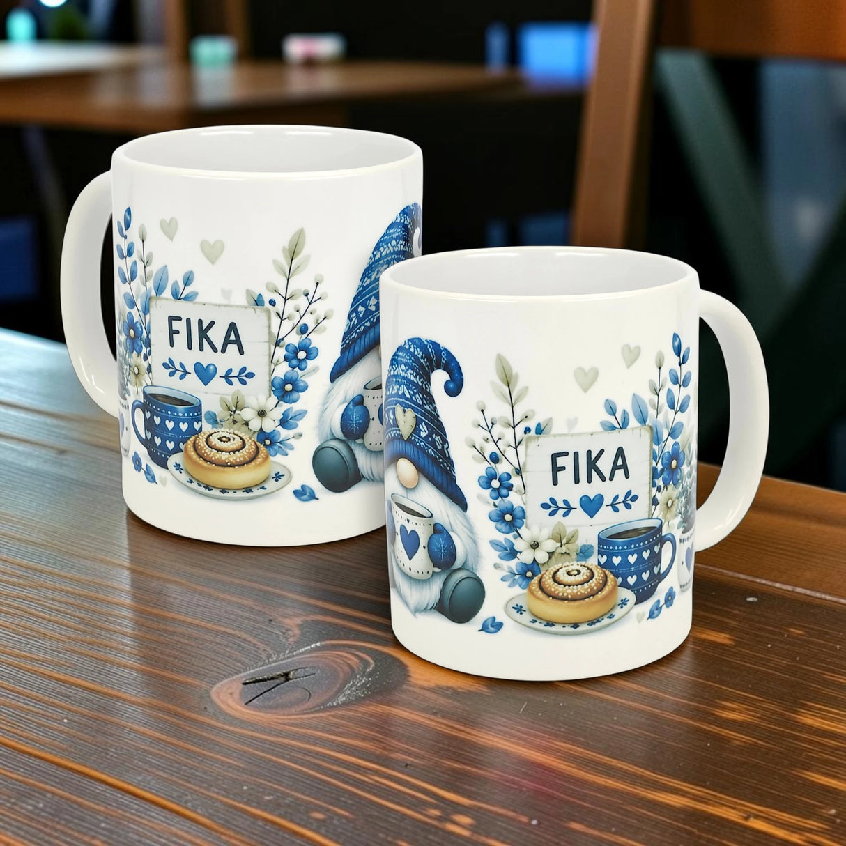 Two Nordic Wholesale Blue Tomte Gnome Fika Ceramic Mugs (11 oz) with blue and gray Scandinavian gnome and foliage designs, plus "FIKA" text, are displayed on a wooden table in a cozy setting.