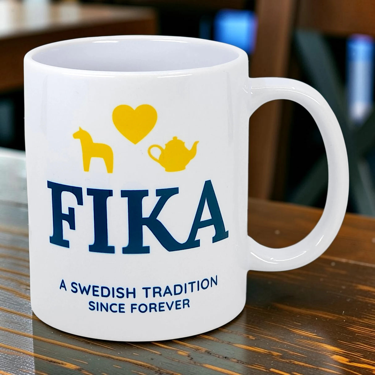 A white ceramic Mug: Fika — A Swedish Tradition Since Forever (11 oz) by Nordic Wholesale rests on a wooden table, featuring the phrase "FIKA: A Swedish tradition since forever" and icons of a yellow horse, heart, and teapot above the text.