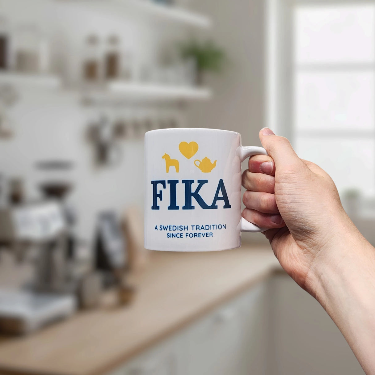 A hand holds the Nordic Wholesale Mug: Fika — A Swedish Tradition Since Forever Ceramic Mug (11 oz), featuring blue and yellow designs, in a blurred kitchen setting.