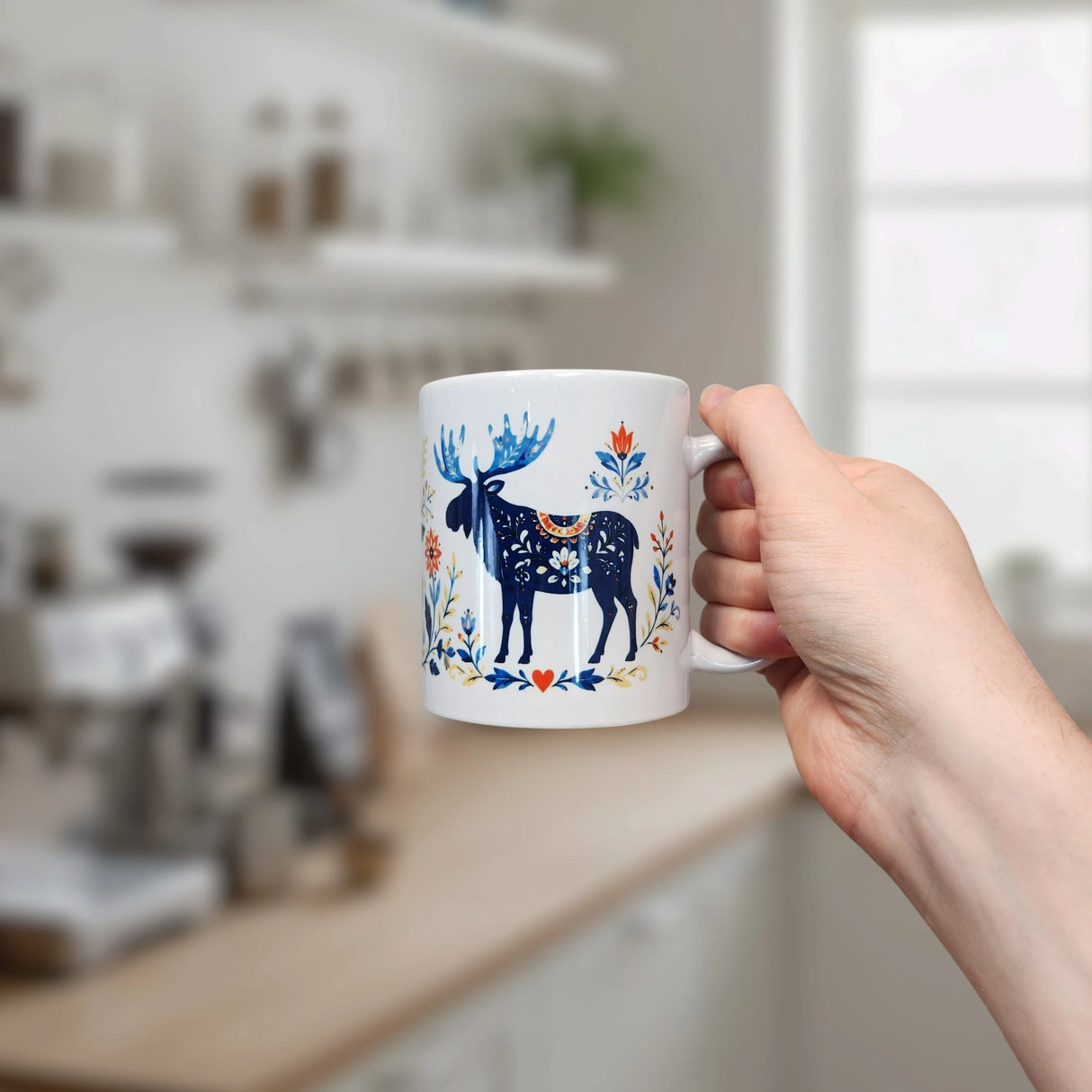 In a bright, modern kitchen, a hand holds the Nordic Wholesale Folk Art Moose Ceramic Mug (11 oz), decorated with blue moose and floral designs; shelves, plants, and kitchenware appear softly blurred in the background.