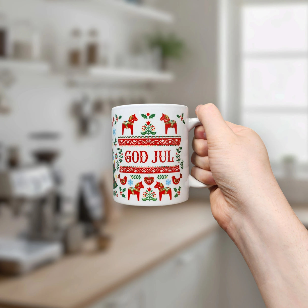A hand holds the Nordic Wholesale Mug: God Jul Dala Horse Christmas Ceramic Mug (11 oz Capacity), a white Scandinavian-style mug decorated with red and green Nordic patterns and the words "God Jul," in a kitchen setting with blurry shelves and a window in the background.