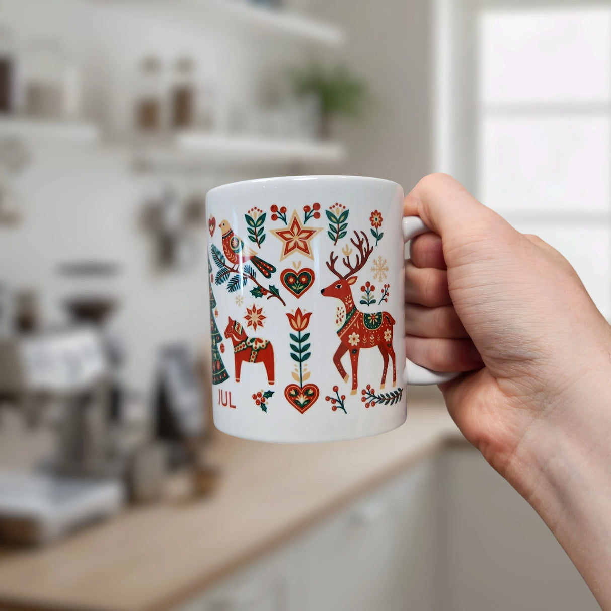 A hand holds the Nordic Wholesale Mug: God Jul Nordic Folk Art Ceramic Mug (11 oz Capacity), featuring colorful Scandinavian-style illustrations of a deer, horse, bird, and hearts, in a softly blurred kitchen setting.