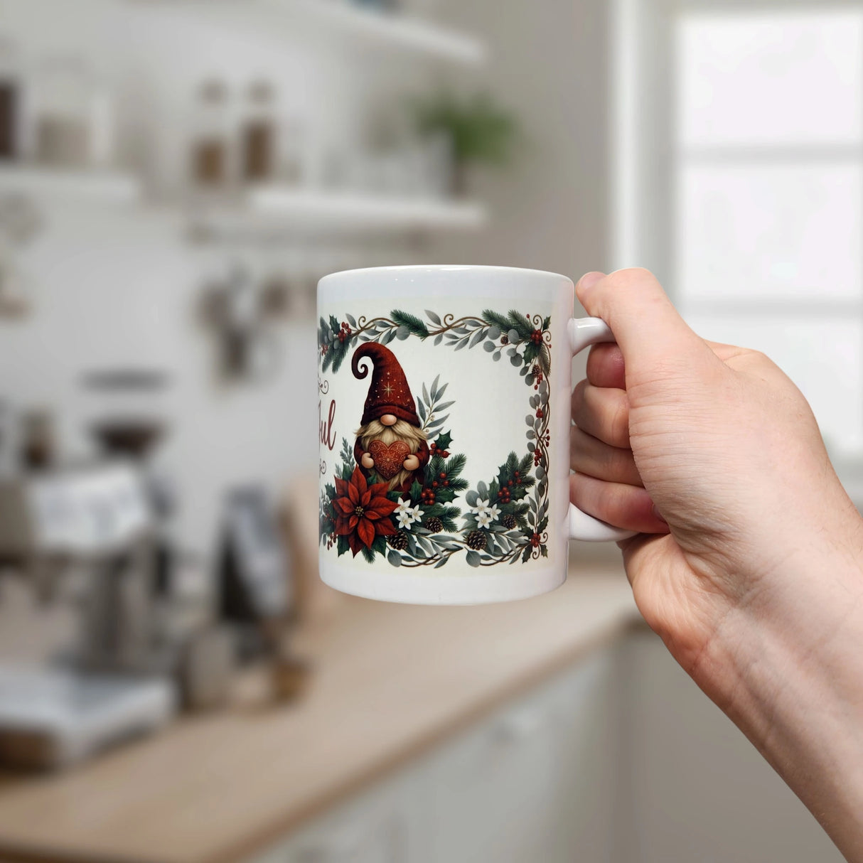 A hand holds the Nordic Wholesale God Jul Tomte Nordic Christmas Ceramic Mug (11 oz), a white Scandinavian-style mug adorned with festive gnomes, poinsettias, and holly. In the background, a softly blurred, light-filled kitchen reveals shelves with jars and a coffee maker.