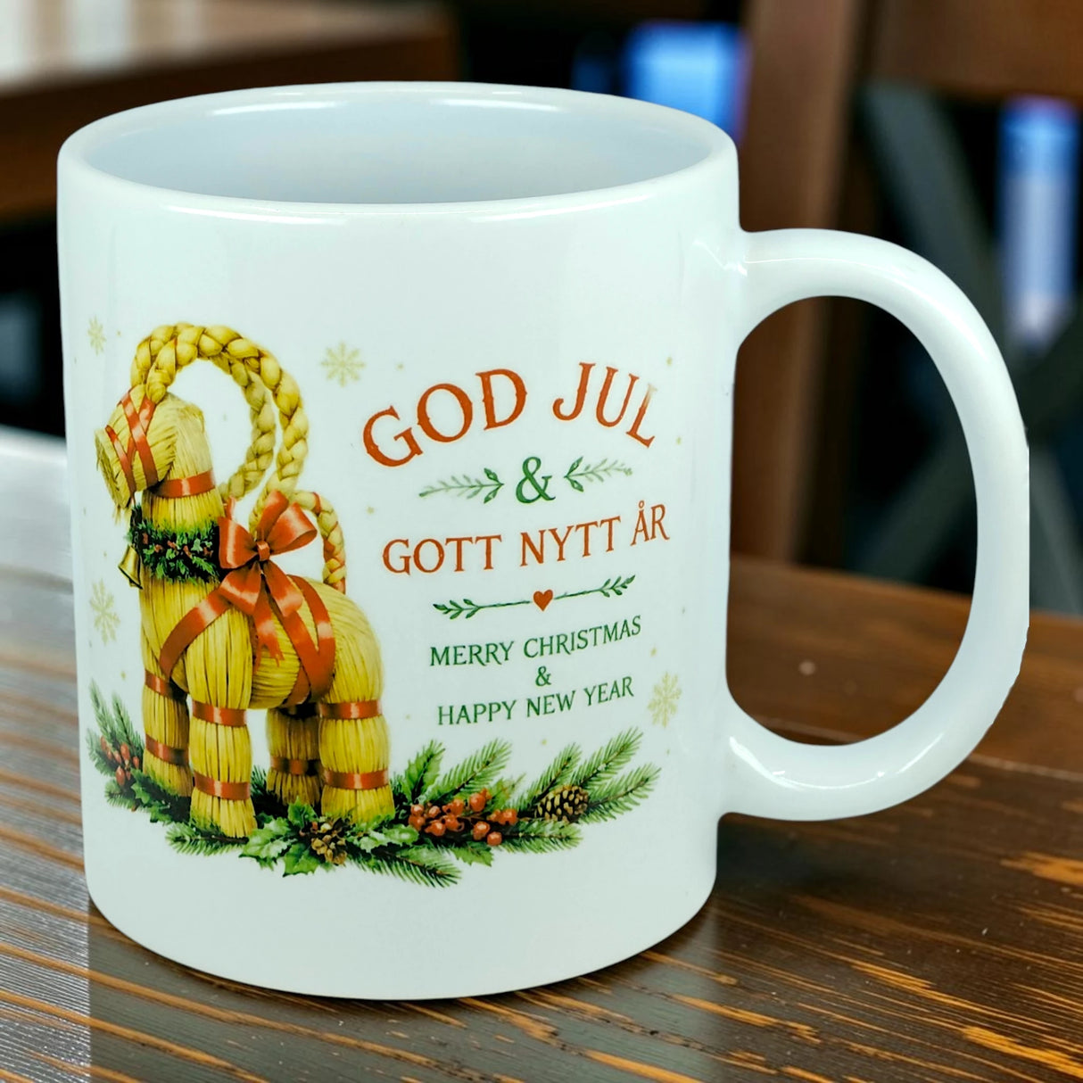 The Nordic Wholesale Mug: God Jul Yule Goat Ceramic Mug (11 oz Capacity) showcases a delightful Scandinavian Yule Goat illustration, pine branches, and the festive text "God Jul & Gott Nytt År — Merry Christmas & Happy New Year." The Swedish Julbock mug is elegantly displayed on a wooden table with a softly blurred background.