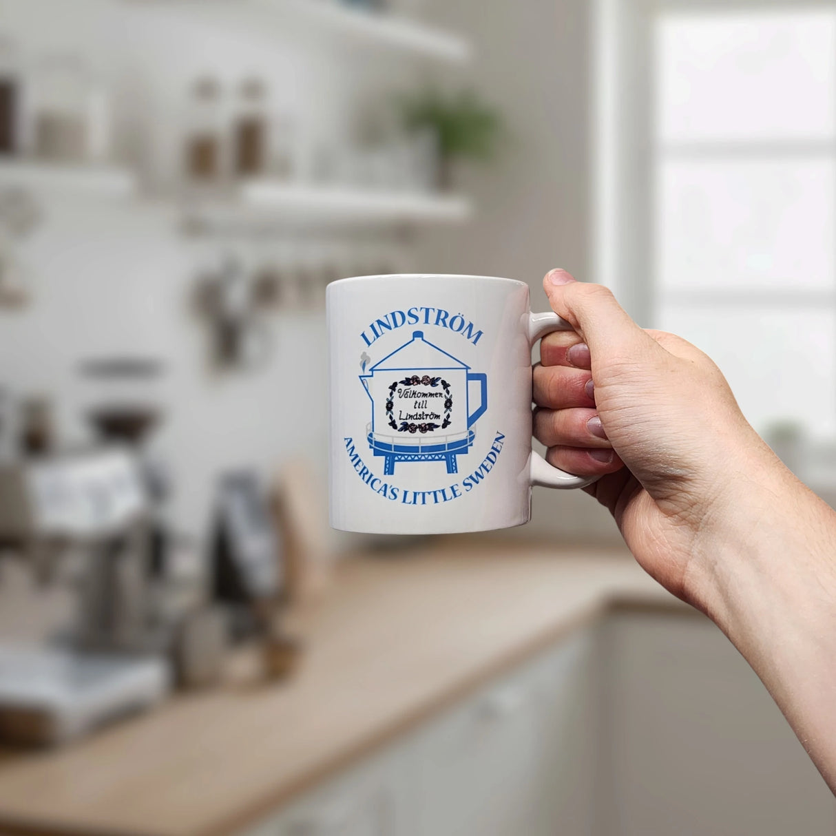 A hand holds the 11 oz ceramic "Lindstrom Coffee Pot Water Tower America's Little Sweden" mug from Swedish Gift Store, featuring blue text and illustration, in a bright kitchen with blurred shelves and window in the background.