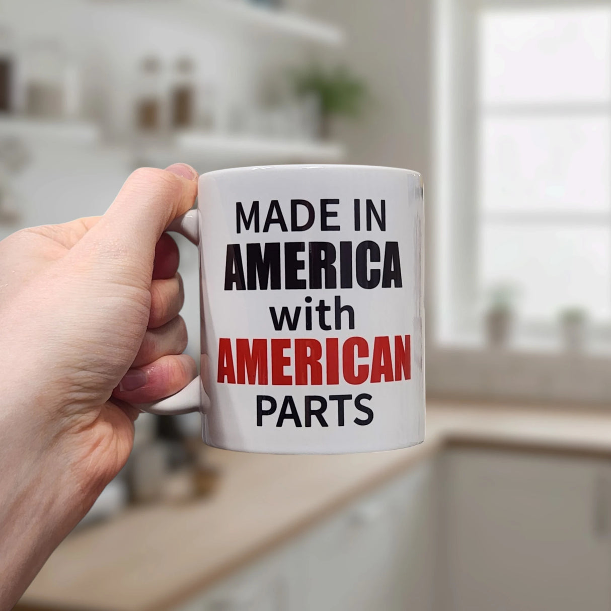 A hand holds the Swedish Gift Store's 11 oz "Made in America with American Parts" ceramic mug in a kitchen. The white mug has bold text—“AMERICAN” in red, other words in black—making it a witty choice for your morning routine.