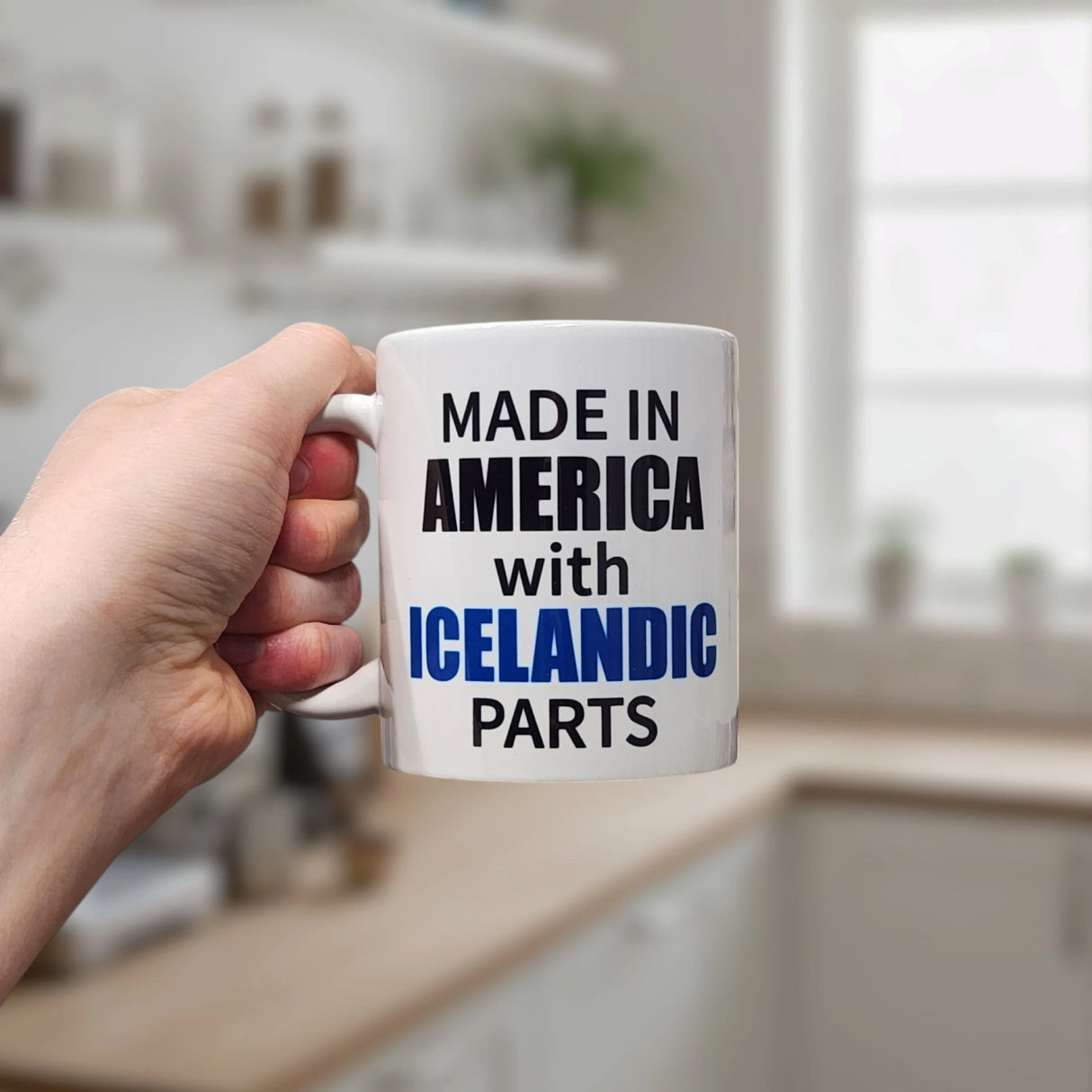 A hand holds a Swedish Gift Store 11 oz ceramic mug in a kitchen. The white mug reads "MADE IN AMERICA with ICELANDIC PARTS" in black and blue text, with blurred shelves and a window visible in the background.