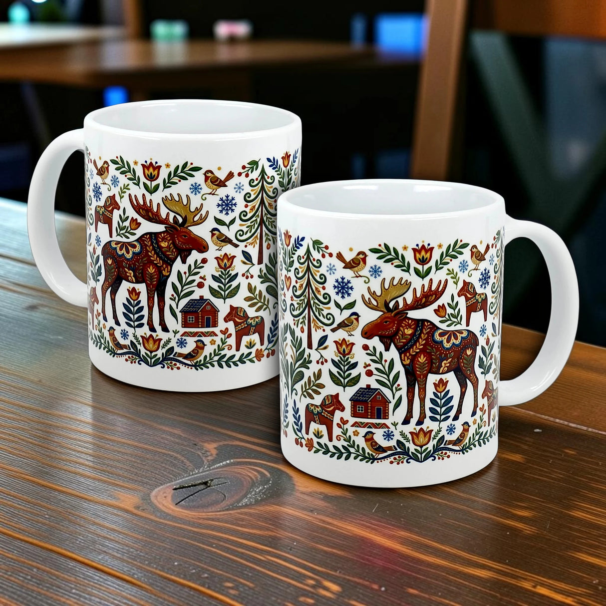 Two white Nordic Forest Kurbits Dala Moose Wraparound Ceramic Mugs (11 oz) by Swedish Gift Store sit on a wooden table, each decorated with Swedish folk-art motifs—moose, trees, houses, and earthy floral patterns.