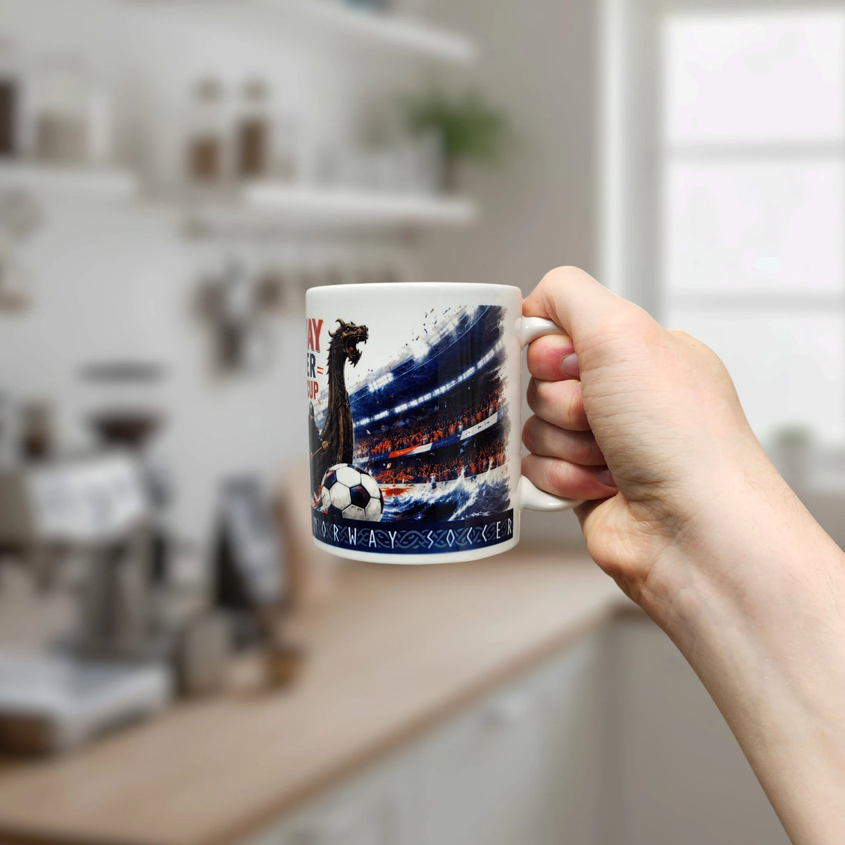 A hand holds the Norway Soccer Coffee Cup Mug – Viking Longship Fan Mug by Swedish Gift Store, featuring a vibrant soccer stadium, a dragon, and a soccer ball, set against a bright, blurred kitchen background.